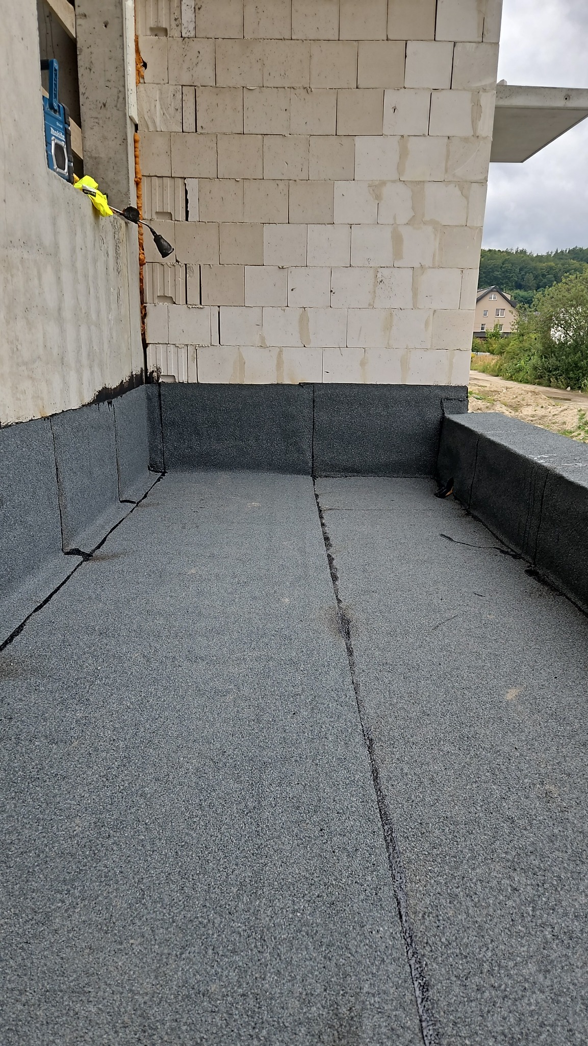 Szczegółowe ujęcie hydroizolacji dachu płaskiego z papy termozgrzewalnej, z widocznymi łączeniami i fragmentem ściany budynku w tle. Praca dekarska w trakcie realizacji.