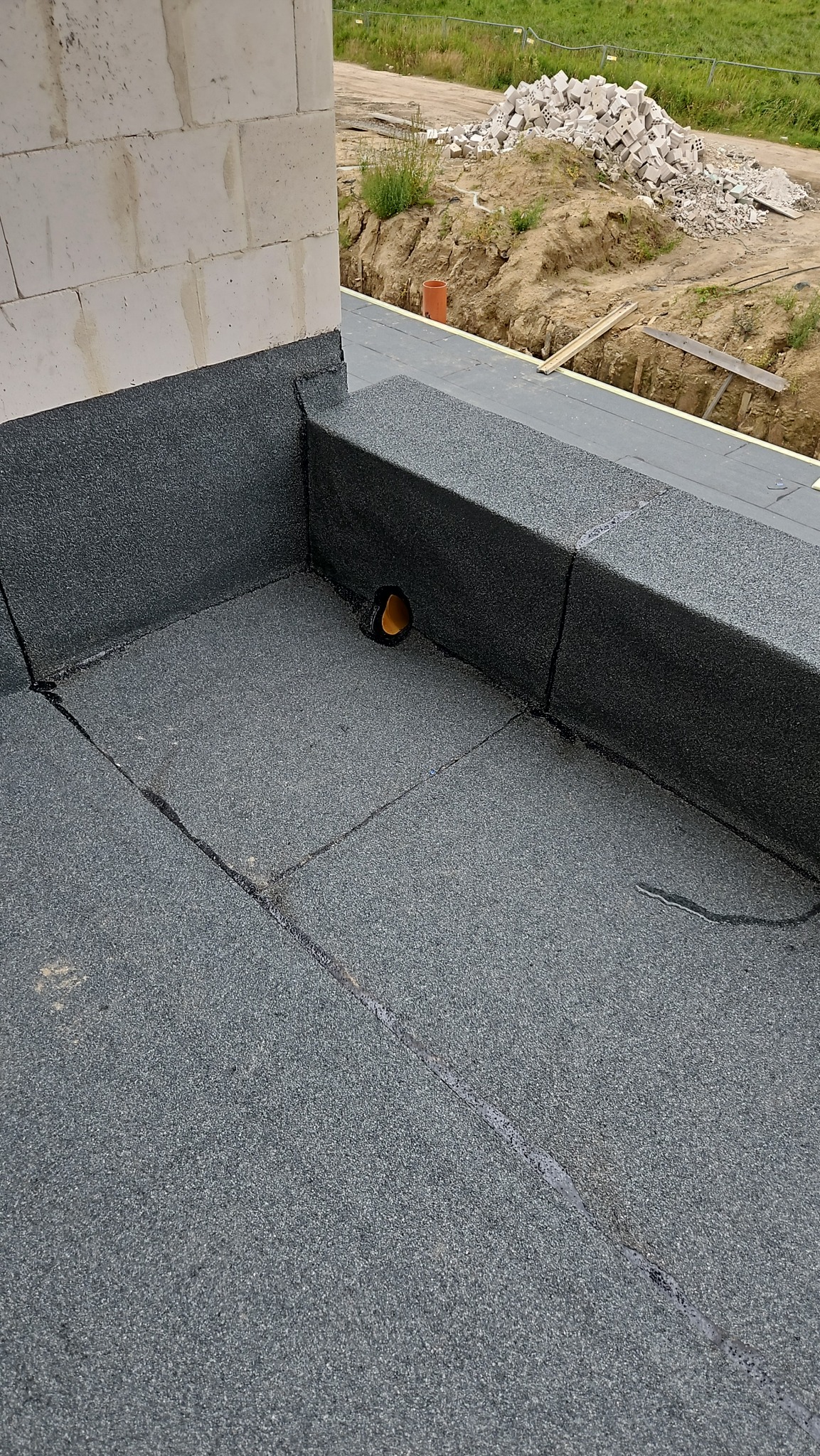 Szczegółowe ujęcie izolacji dachu płaskiego z papy termozgrzewalnej, z widocznym odpływem wody i fragmentem ściany z betonu komórkowego w tle.