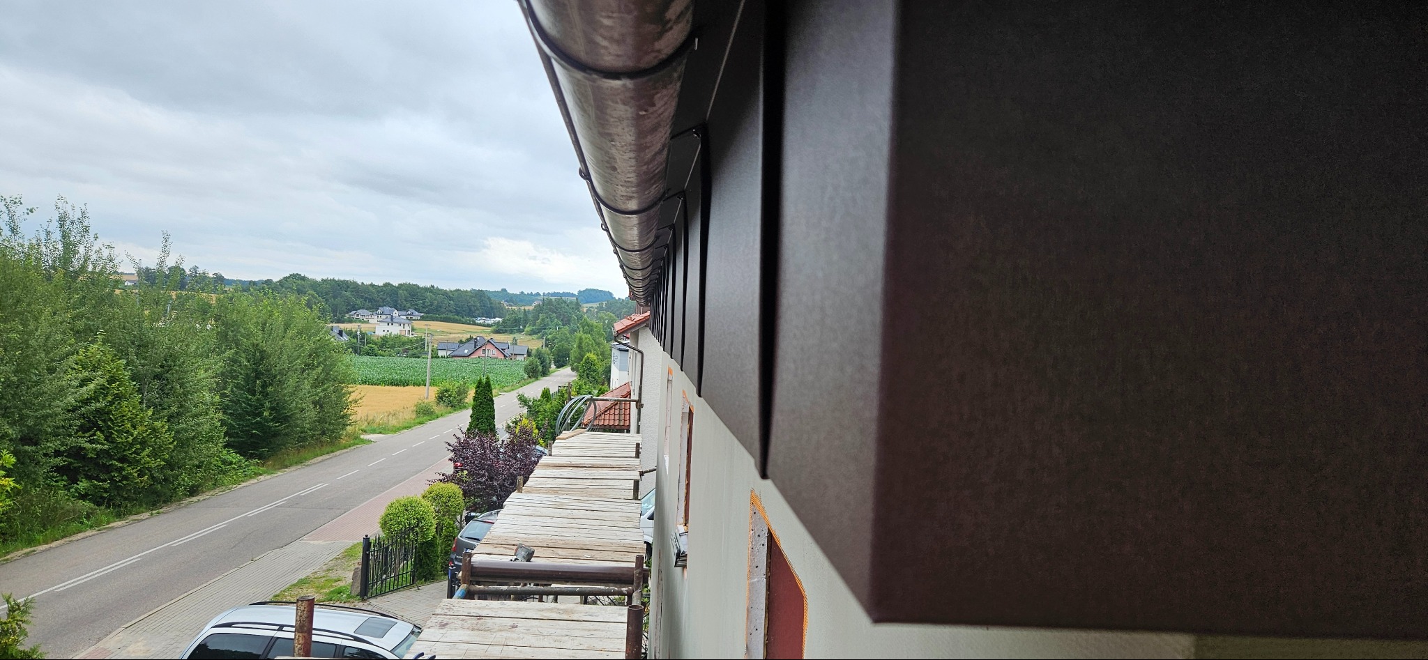 Zamontowany system rynnowy w kolorze grafitowym, widok z rusztowania na tle krajobrazu wiejskiego. Detal wykończenia dachu z bliska.