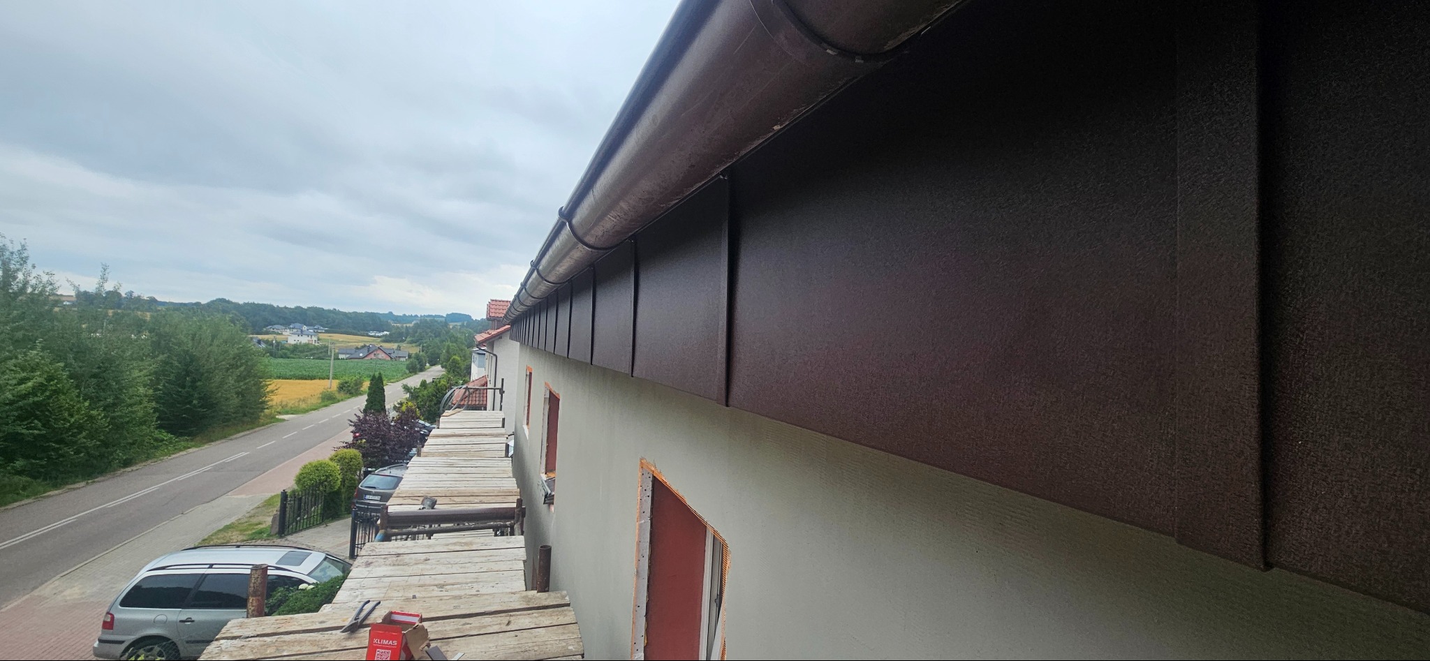 Zamontowana, brązowa podbitka dachowa z rynną, widoczna z rusztowania na tle krajobrazu. Detal wykończenia dachu, estetyczne połączenie z elewacją budynku.