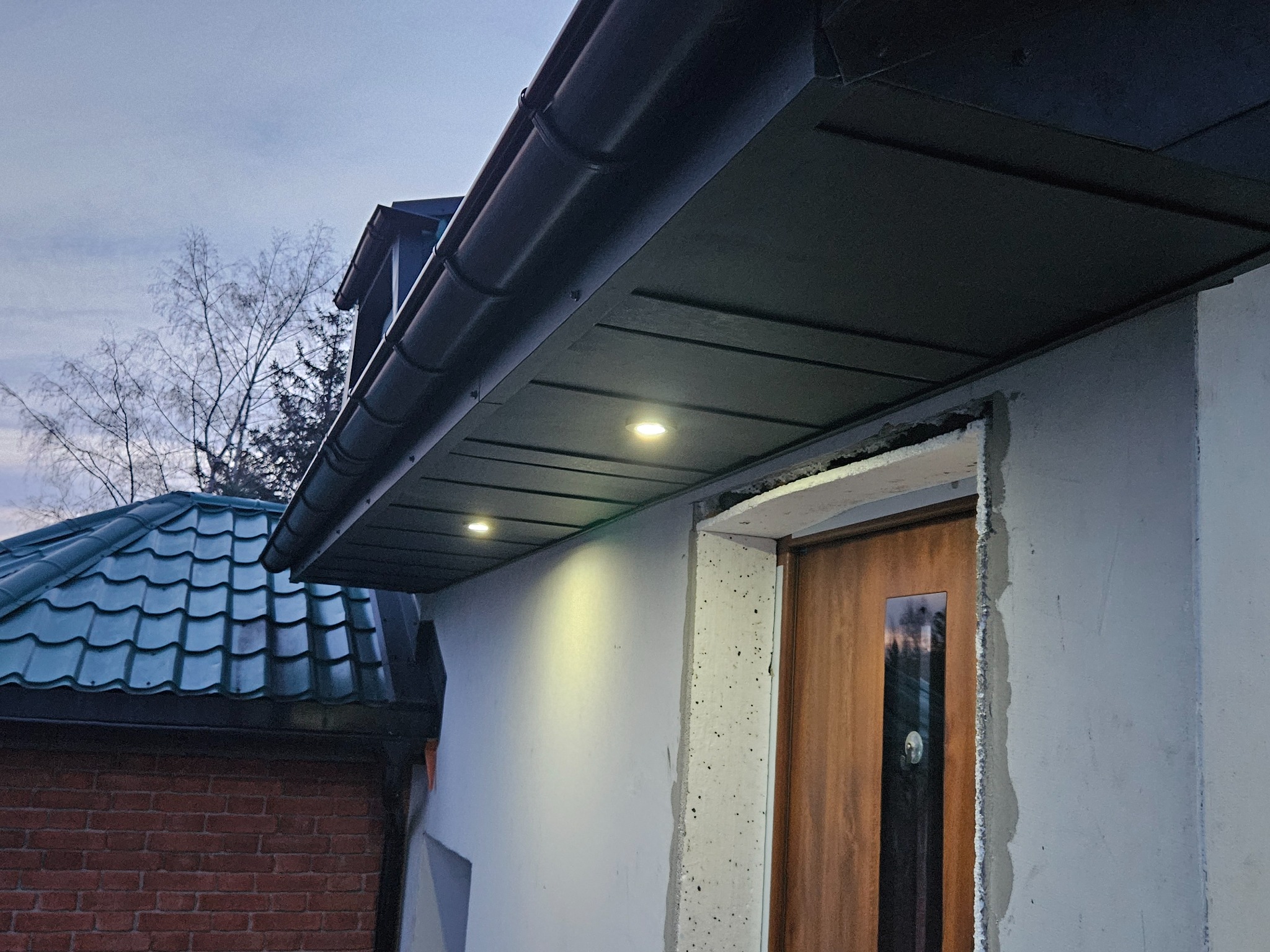 Nowoczesne podbitka dachowa z oświetleniem punktowym, widok z dołu na fragment dachu i elewacji budynku z drzwiami wejściowymi. Dachówka w tle.