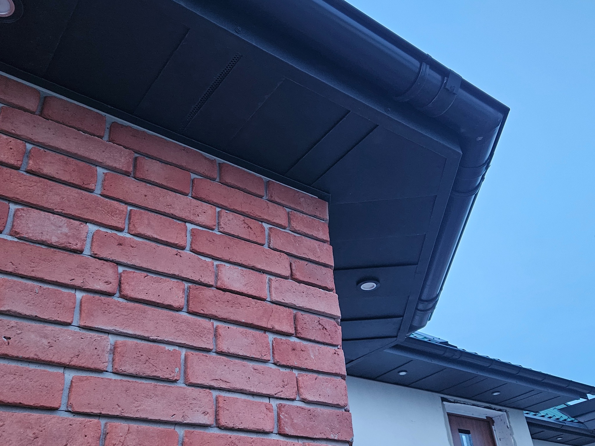 Ceglany narożnik budynku z ciemną podbitką dachową i rynną w zbliżeniu, widoczne oświetlenie punktowe. Ujęcie z dołu, błękitne niebo w tle.