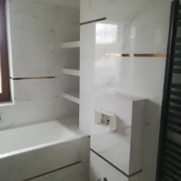 Nowoczesna łazienka w trakcie wykańczania: białe marmurowe płytki z akcentami w kolorze miedzi, zabudowana wanna, półki ścienne i grzejnik drabinkowy. Widoczna puszka instalacyjna podtynkowego WC...