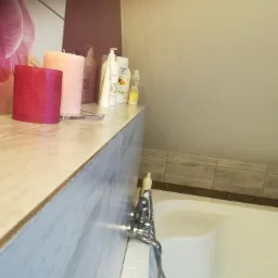 Ustawione na blacie łazienkowym świece i kosmetyki, w tle wanna i kwiatowy motyw dekoracyjny na ścianie.