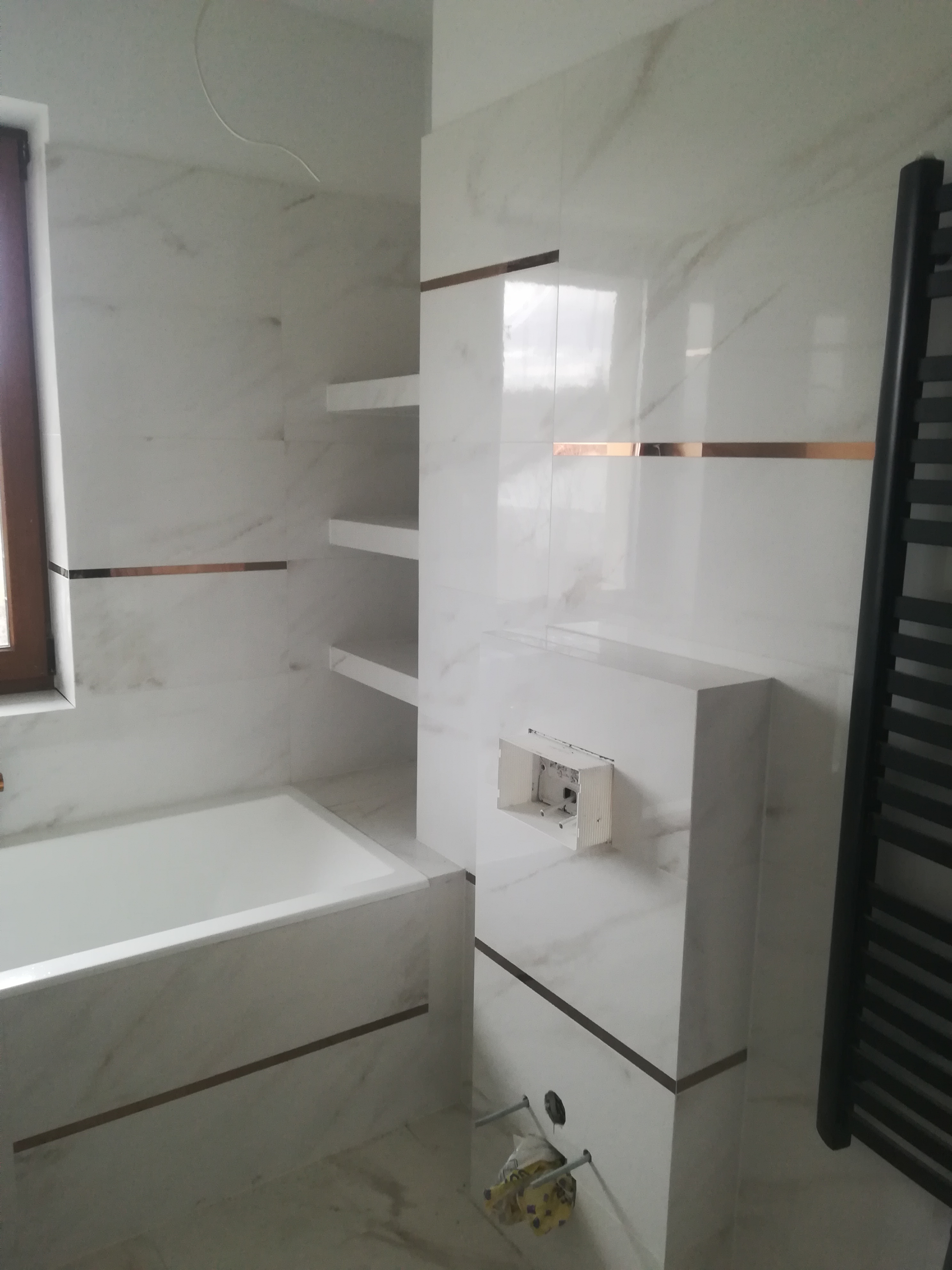 Nowoczesna łazienka w trakcie wykańczania: białe marmurowe płytki z akcentami w kolorze miedzi, zabudowana wanna, półki ścienne i grzejnik drabinkowy. Widoczna puszka instalacyjna podtynkowego WC...