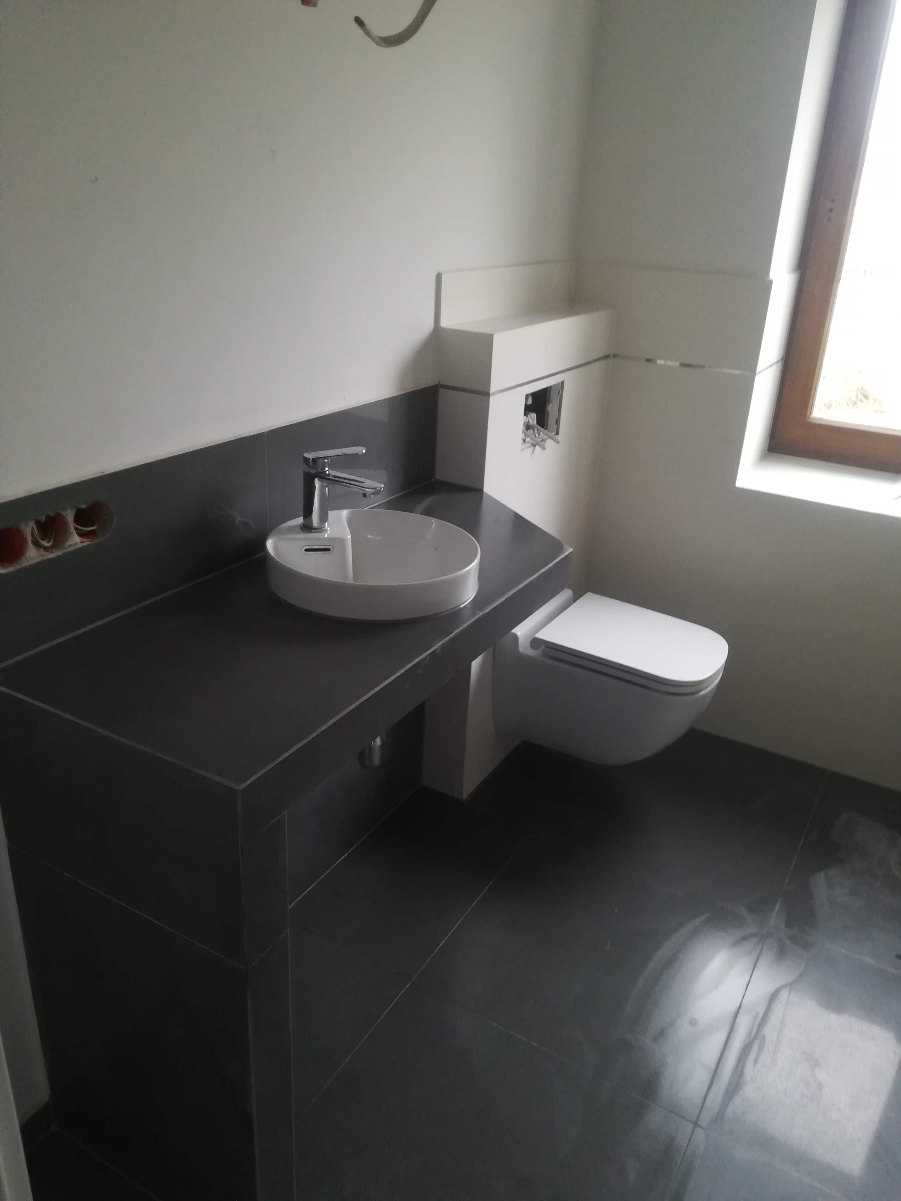 Nowoczesna łazienka z wiszącą toaletą i umywalką nablatową na ciemnoszarym blacie, widoczne niezabezpieczone przewody elektryczne w ścianie.