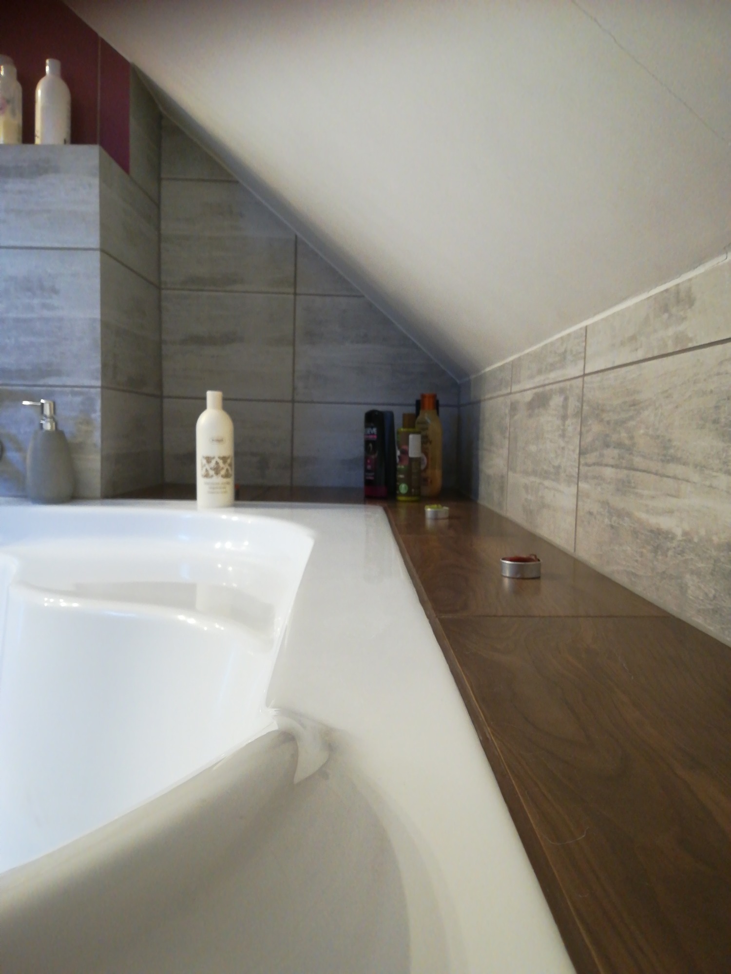 Biała wanna w łazience na poddaszu, z drewnianym blatem, szarymi płytkami imitującymi drewno i kolekcją kosmetyków.