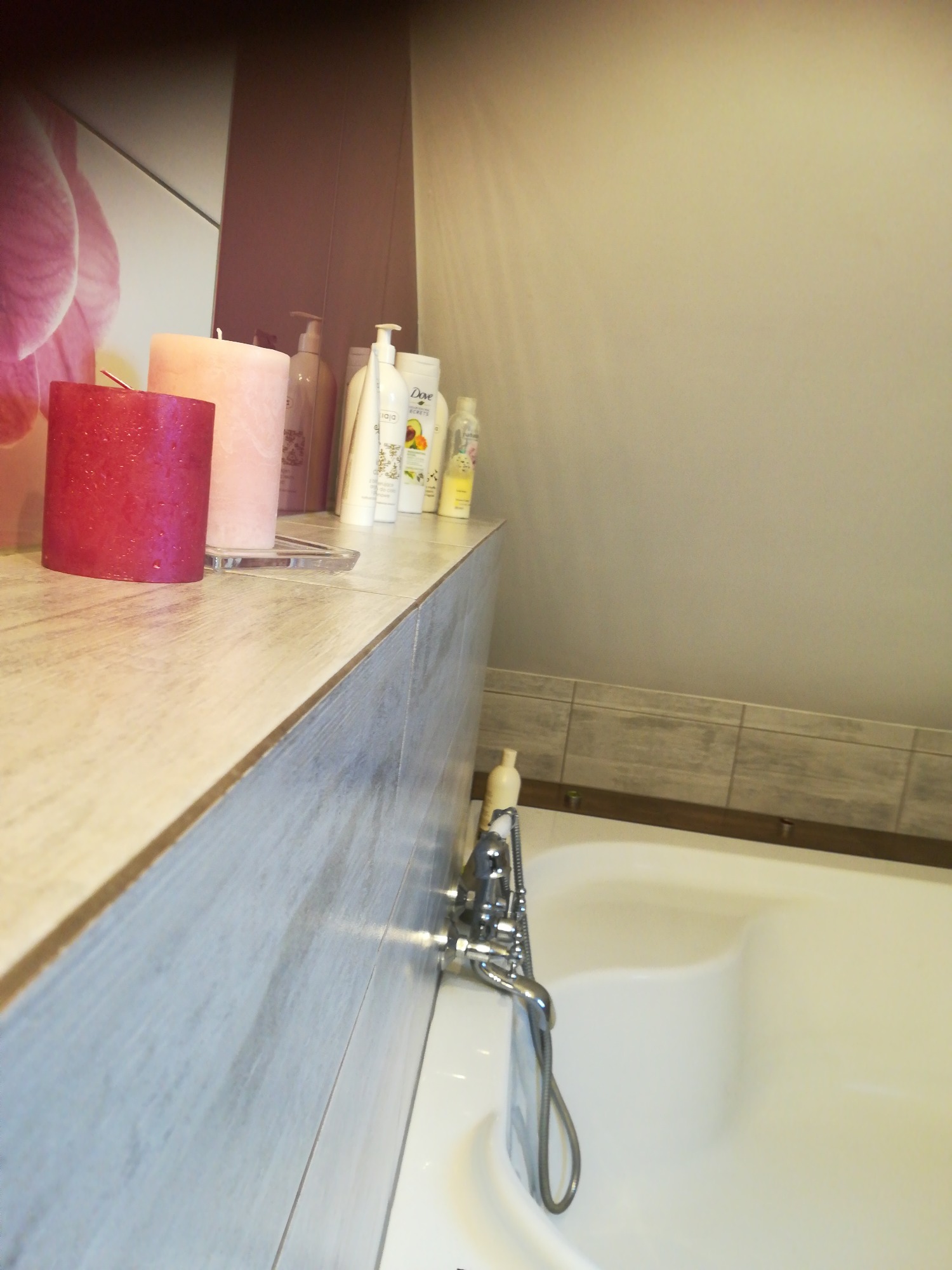 Ustawione na blacie łazienkowym świece i kosmetyki, w tle wanna i kwiatowy motyw dekoracyjny na ścianie.