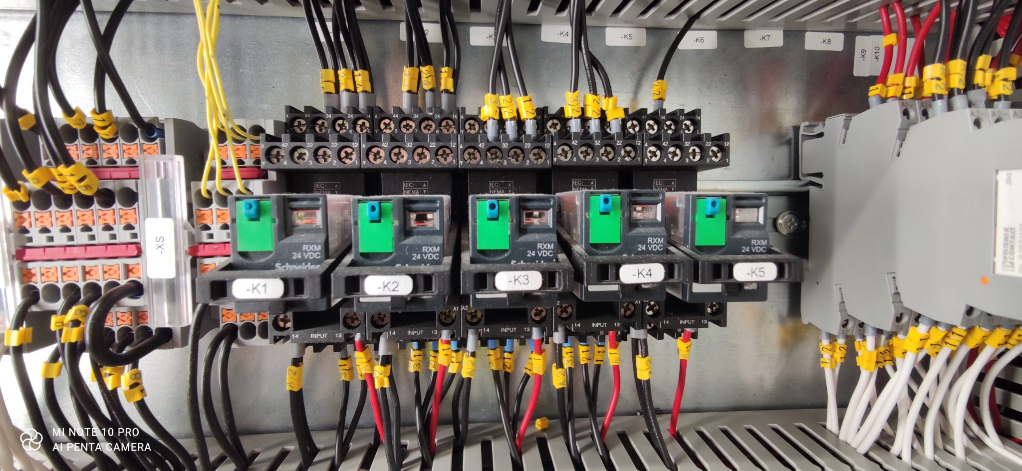 Szczegółowe ujęcie panelu sterowania z przekaźnikami, okablowaniem i oznaczeniami, widoczne przekaźniki Schneider RXM 24 VDC z zielonymi wskaźnikami, czarne i kolorowe przewody z żółtymi...