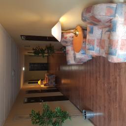 Paweł Markowski - Długi korytarz z drewnianą podłogą, wzorzystymi fotelami, lampkami nocnymi i doniczkowymi roślinami w budynku na wynajem.