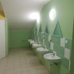 Rząd umywalek w publicznej toalecie z zielonymi ścianami i trójkątnymi lustrami, oświetlonych okrągłymi lampami, z podłogą wyłożoną kafelkami.