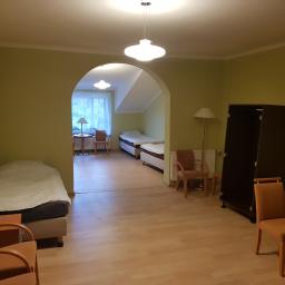 Paweł Markowski - Pokój z łóżkami, szafą i krzesłami, widok przez łukowe przejście do drugiego pokoju, oświetlony lampami i światłem dziennym.