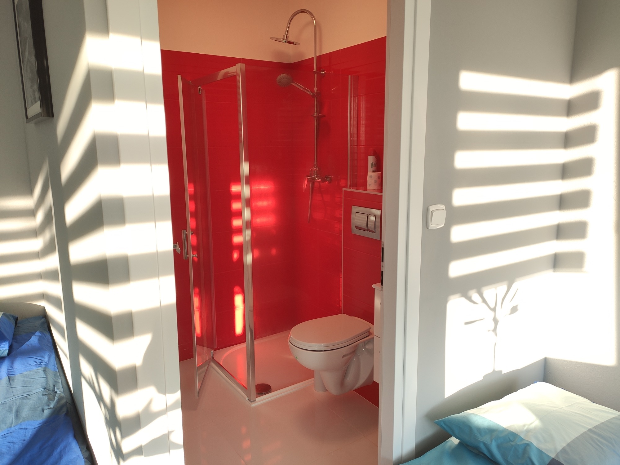 Nowoczesna łazienka z czerwoną kabiną prysznicową, białą toaletą i grzejnikiem, oświetlona promieniami słonecznymi wpadającymi przez żaluzje.