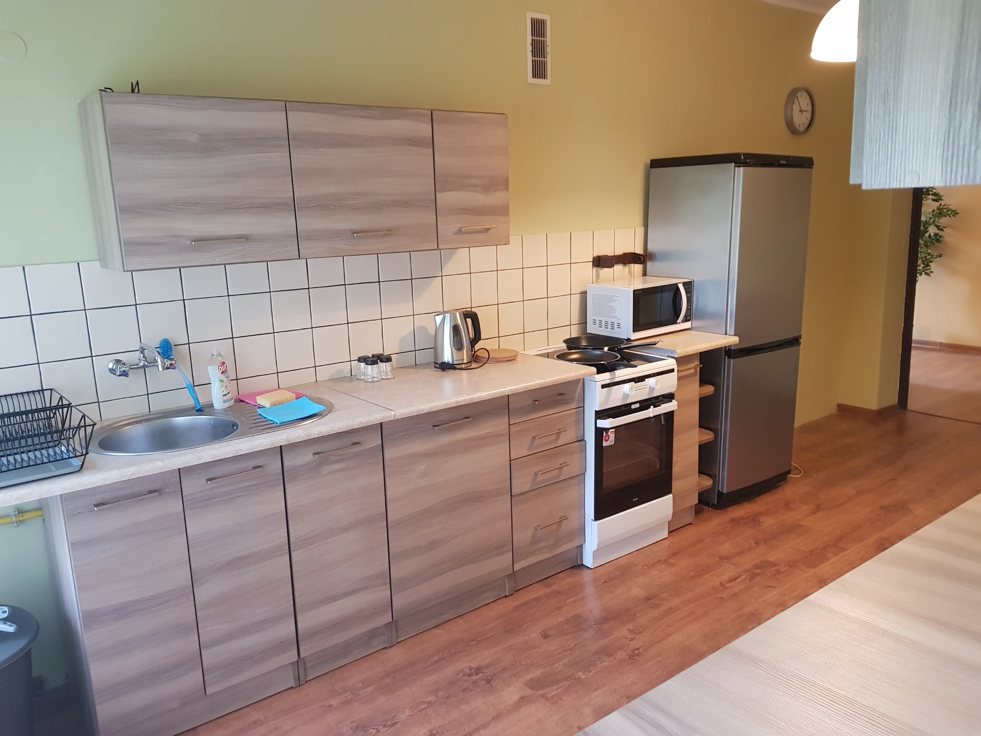 Jasna kuchnia z drewnianymi szafkami, białymi płytkami i srebrną lodówką, widok na otwarte przejście do innego pomieszczenia.