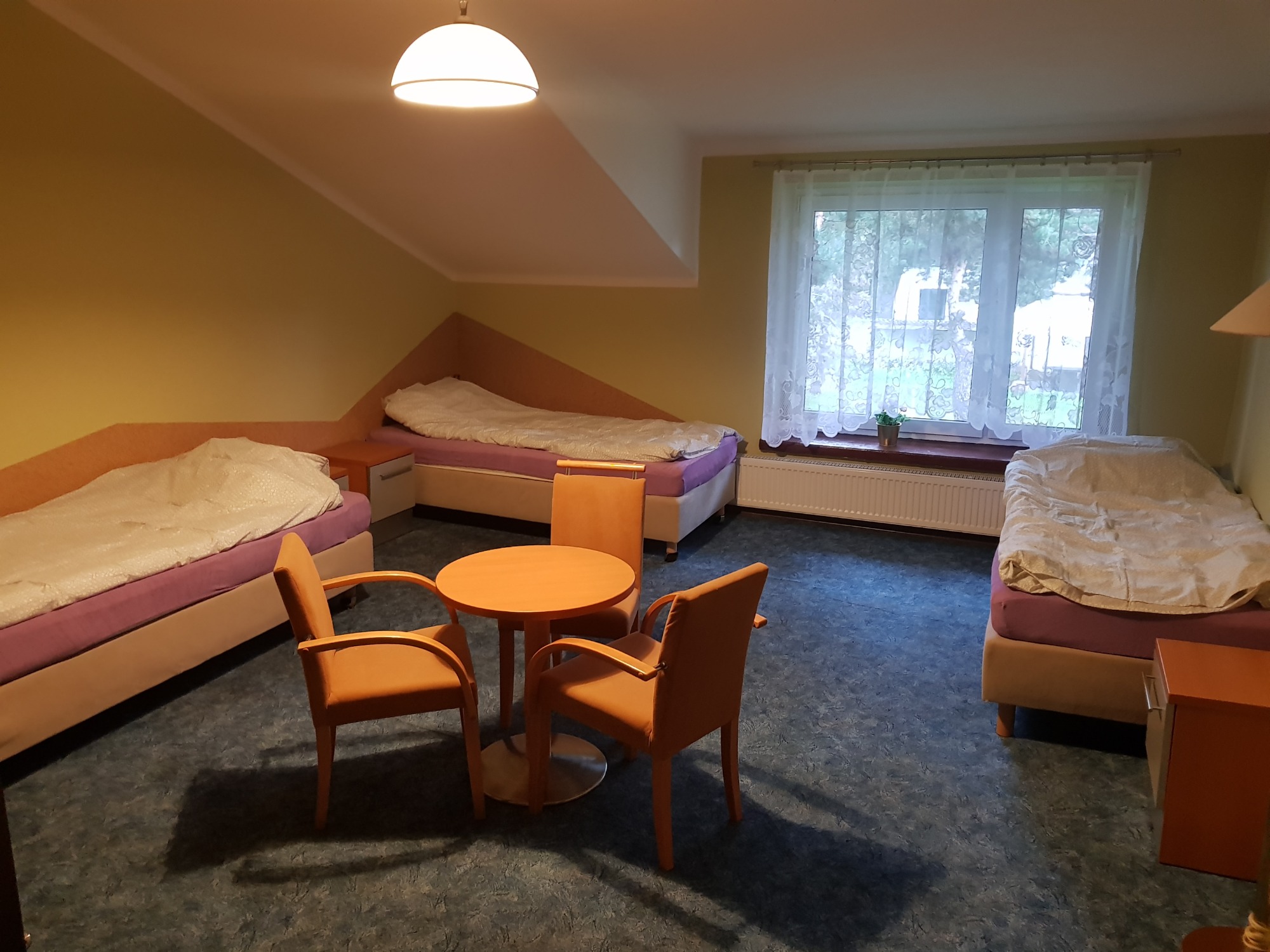 Wnętrze pokoju gościnnego w Biskupicach z trzema łóżkami, okrągłym stolikiem i krzesłami. Widok na pokój z lekkiego podwyższenia, ukazujący ukośne ściany poddasza i okno z firanką.