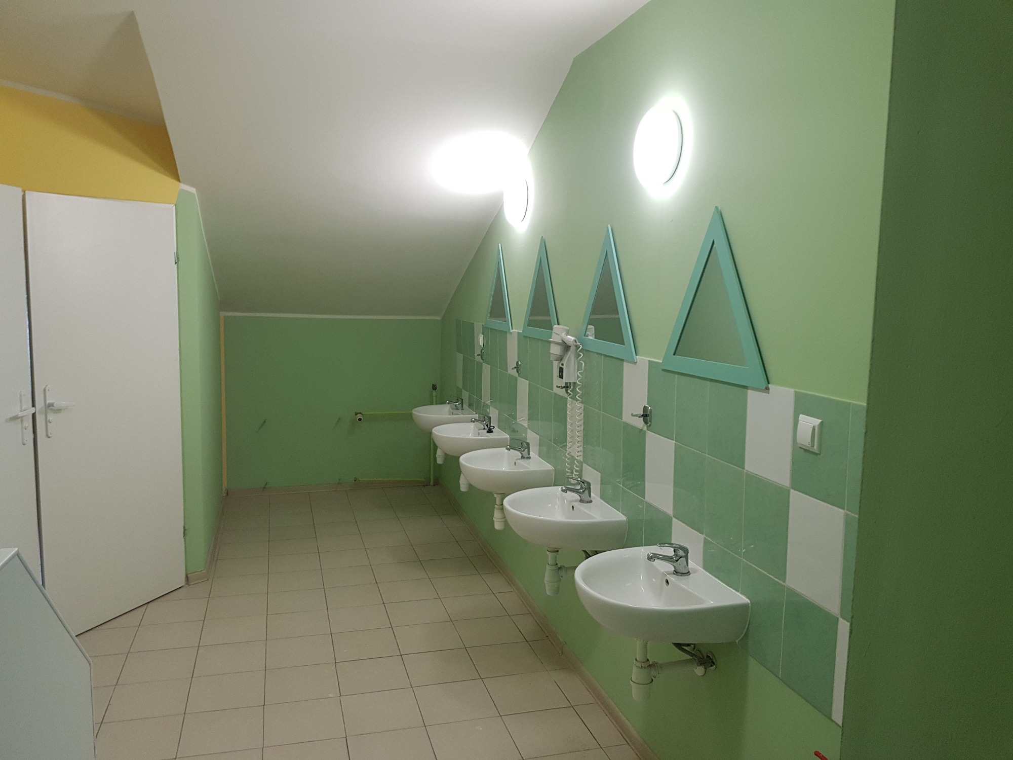 Rząd umywalek w publicznej toalecie z zielonymi ścianami i trójkątnymi lustrami, oświetlonych okrągłymi lampami, z podłogą wyłożoną kafelkami.