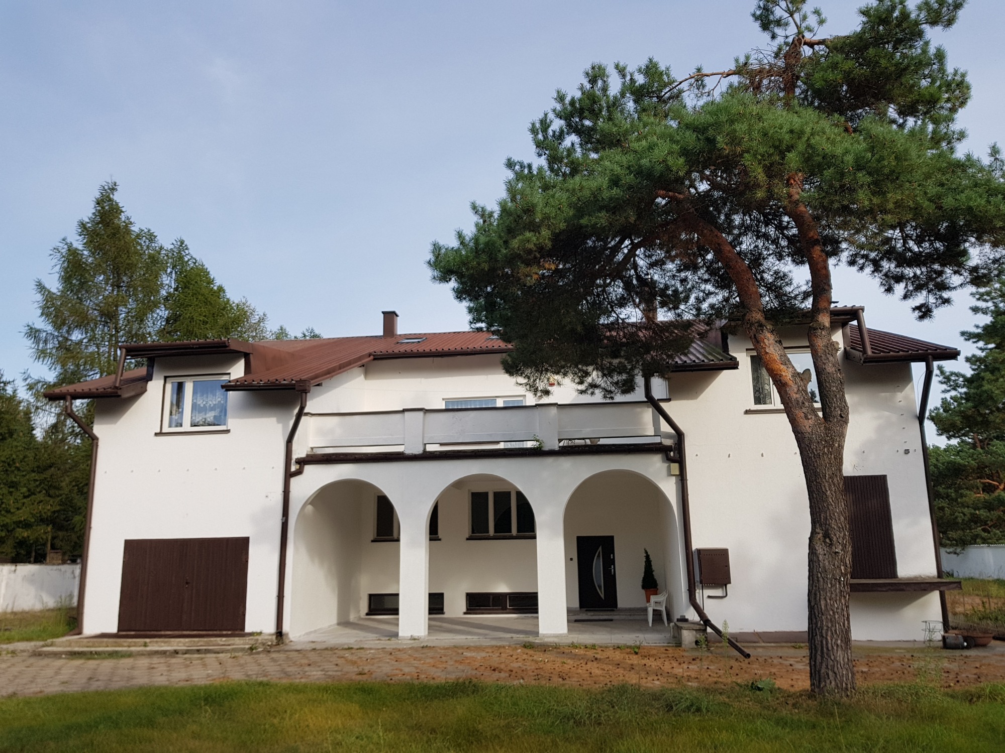 Biały dom z brązowym dachem i garażem, widoczny podjazd z arkadami i duża sosna przed budynkiem.