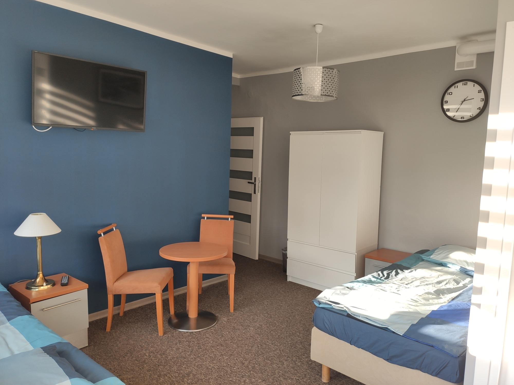 Pokój hotelowy z dwoma łóżkami, stolikiem i krzesłami, telewizorem na ścianie i zegarem. Widoczna szafa i lampka nocna.