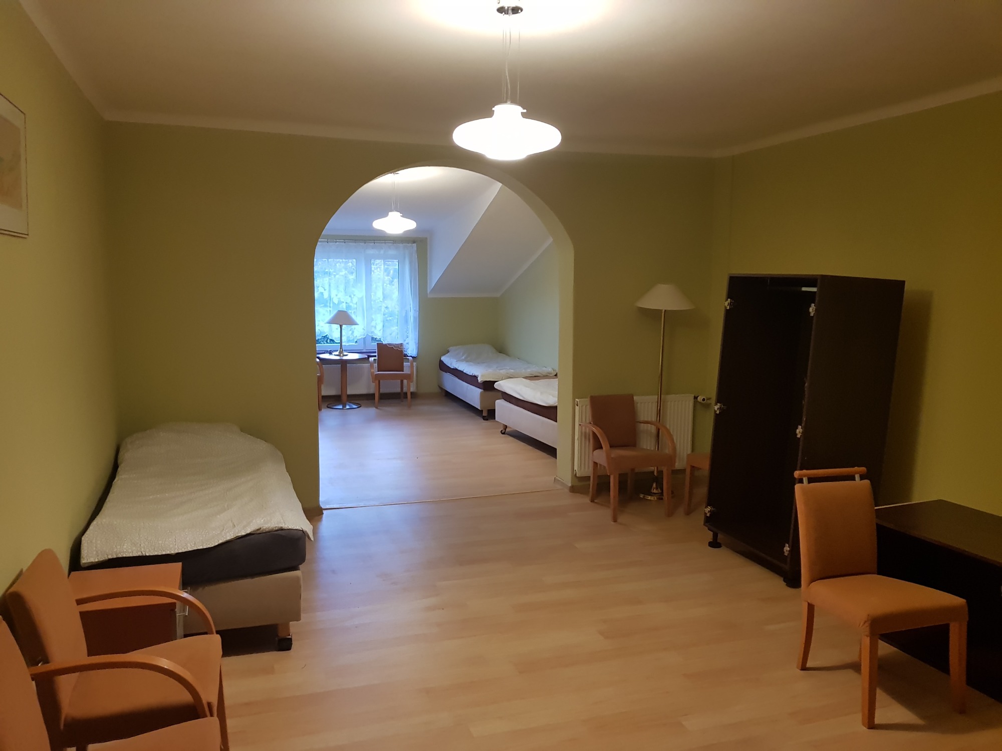 Pokój z łóżkami, szafą i krzesłami, widok przez łukowe przejście do drugiego pokoju, oświetlony lampami i światłem dziennym.