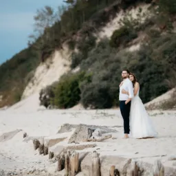 Para młoda, mężczyzna w białej koszuli i ciemnych spodniach, kobieta w białej sukni ślubnej, obejmują się na piaszczystej plaży z widokiem na zalesioną wydmę.