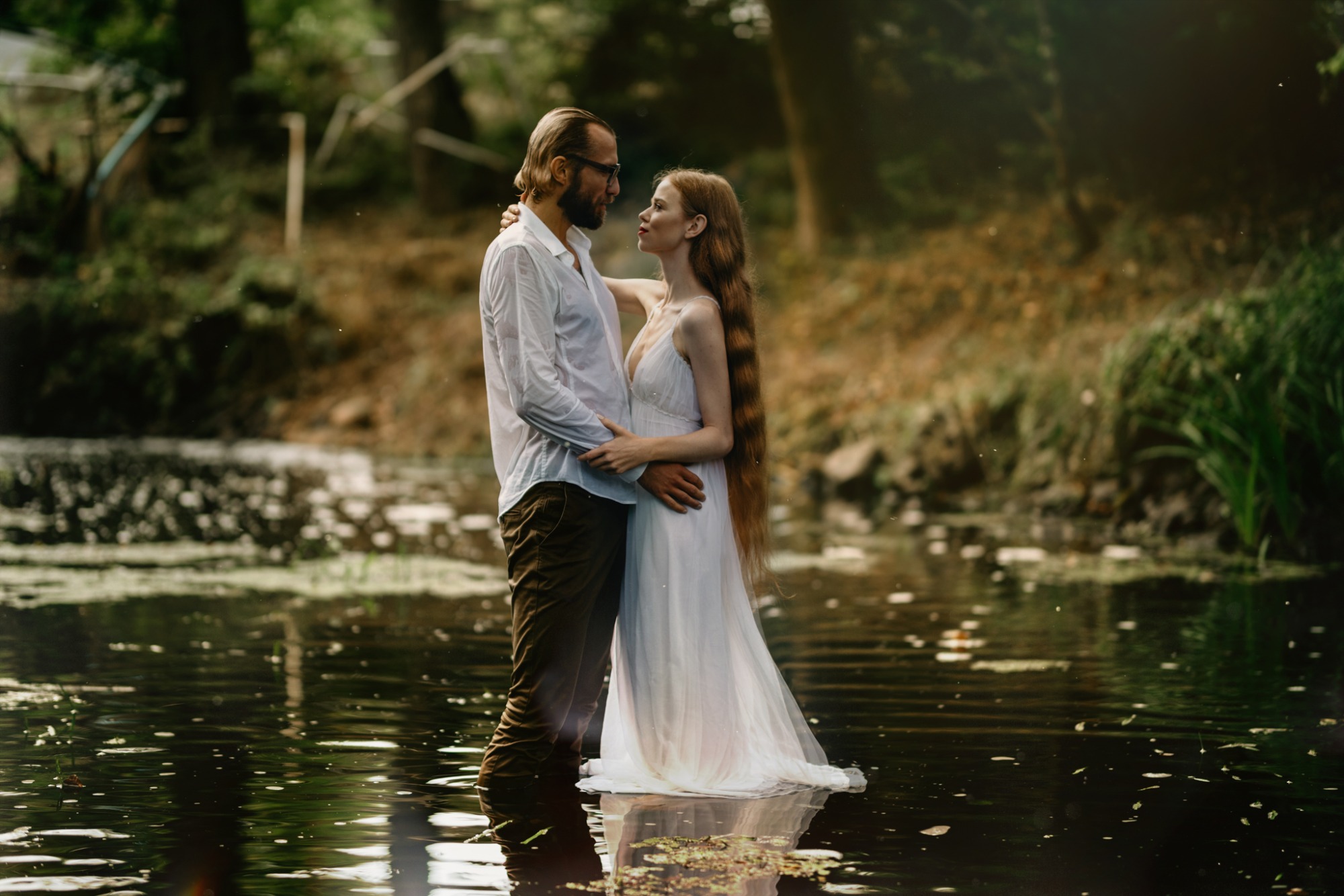 Zakochana para w wodzie, kobieta w białej sukni, mężczyzna w rozpiętej koszuli, romantyczna sceneria leśnego stawu.