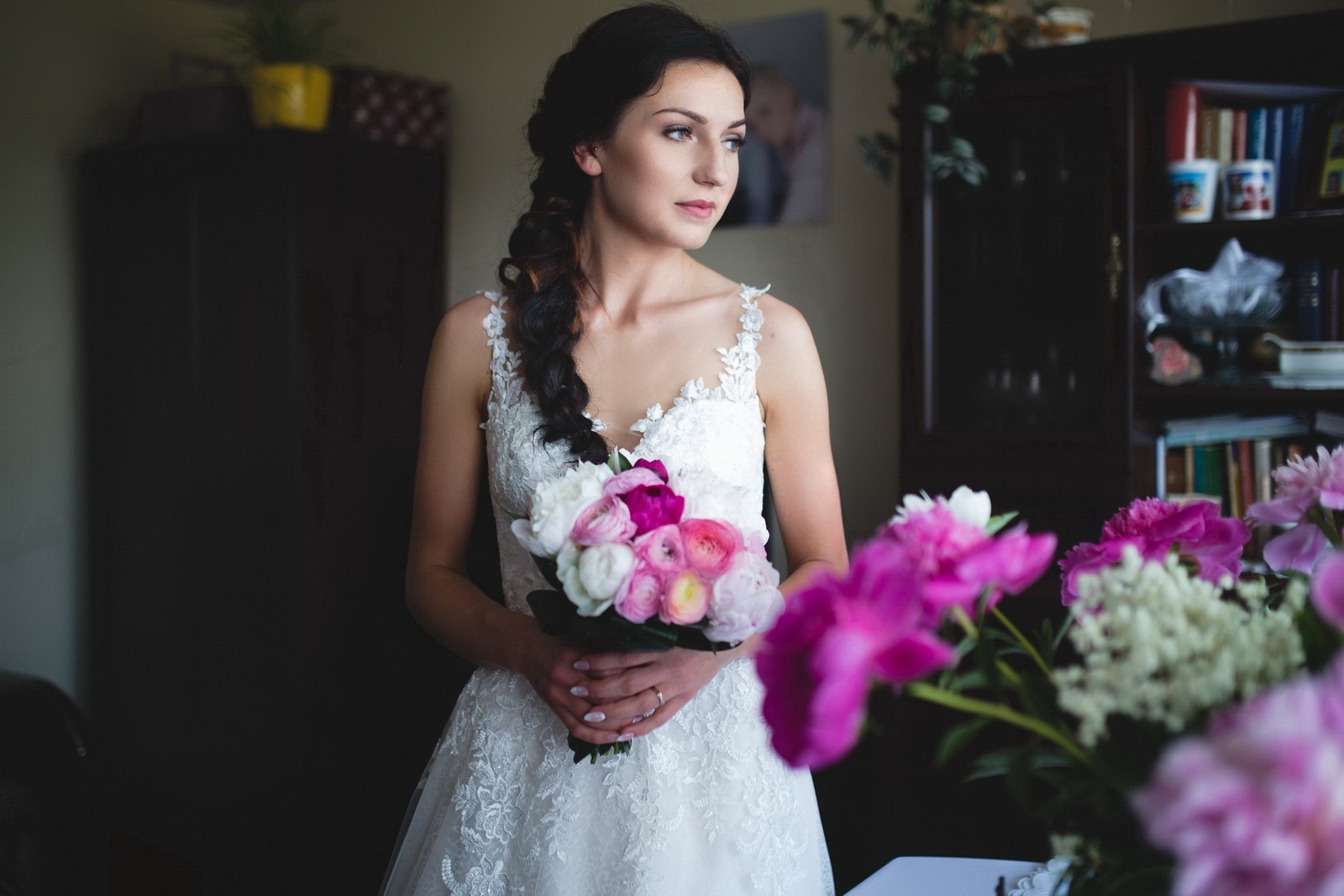 Panna młoda w koronkowej sukni, z warkoczem i bukietem różowych i białych kwiatów, spogląda w bok, w tle ciemne meble i książki.