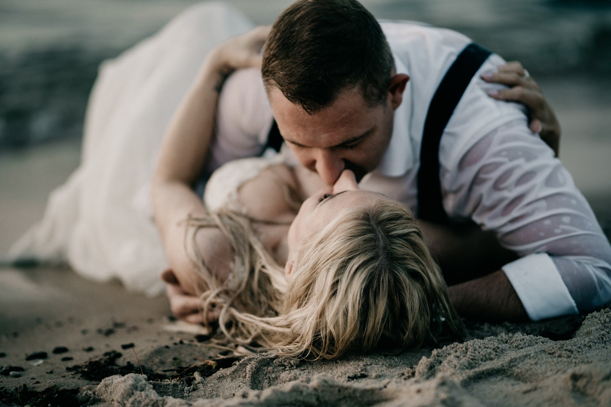 Mokra para młoda w objęciach na plaży, panna młoda leży na piasku, a pan młody opiera się nad nią, oboje mają mokre ubrania.