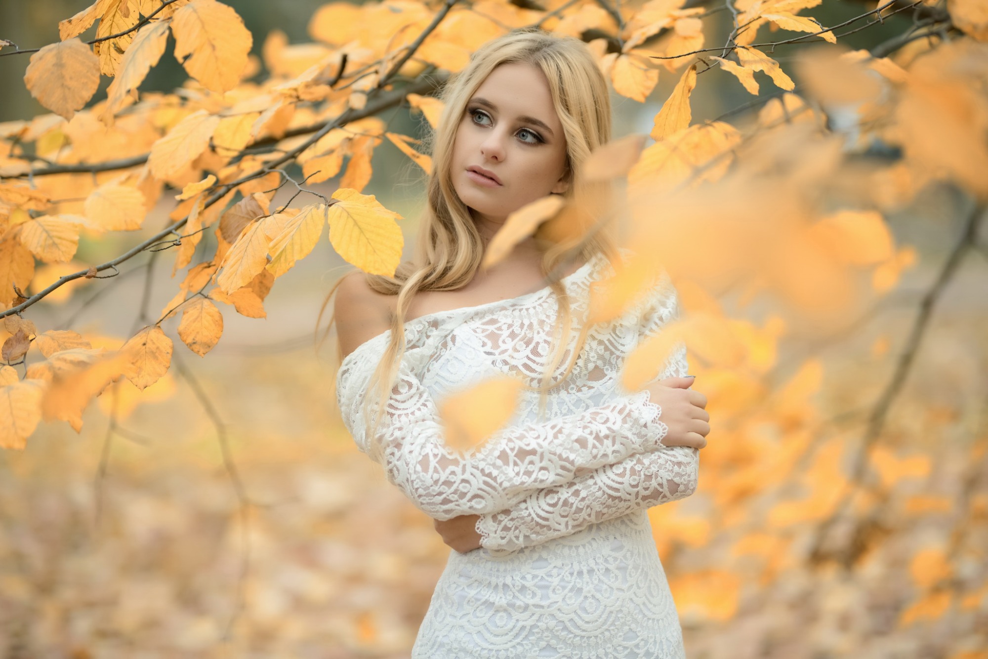 Młoda kobieta w koronkowej sukience pozuje na tle jesiennych liści, zamyślona spogląda w bok.