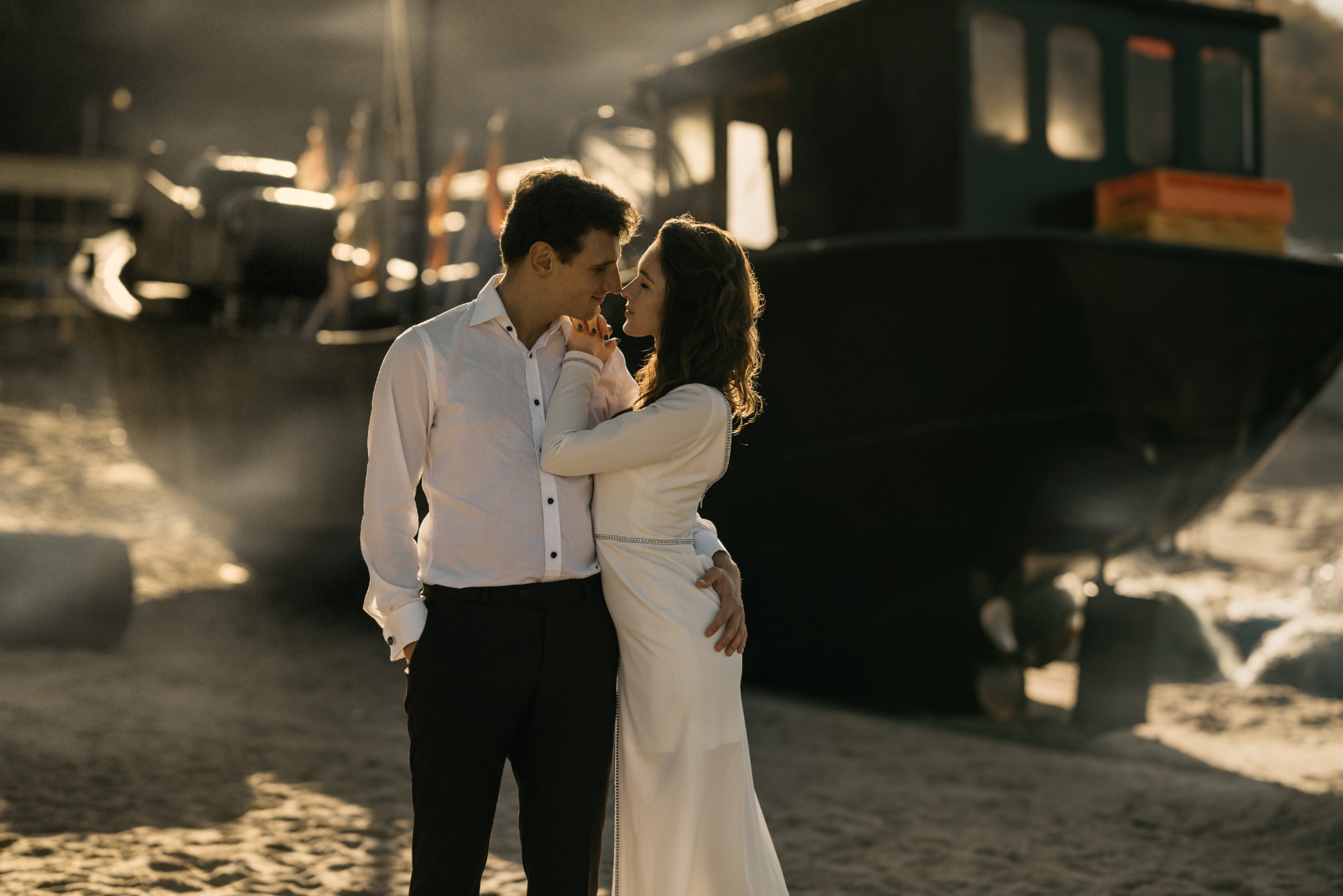 Para narzeczonych, ona w białej sukni z długim rękawem, on w białej koszuli i czarnych spodniach, obejmująca się na plaży z ciemną łodzią rybacką w tle, w ciepłym, zachodzącym słońcu.