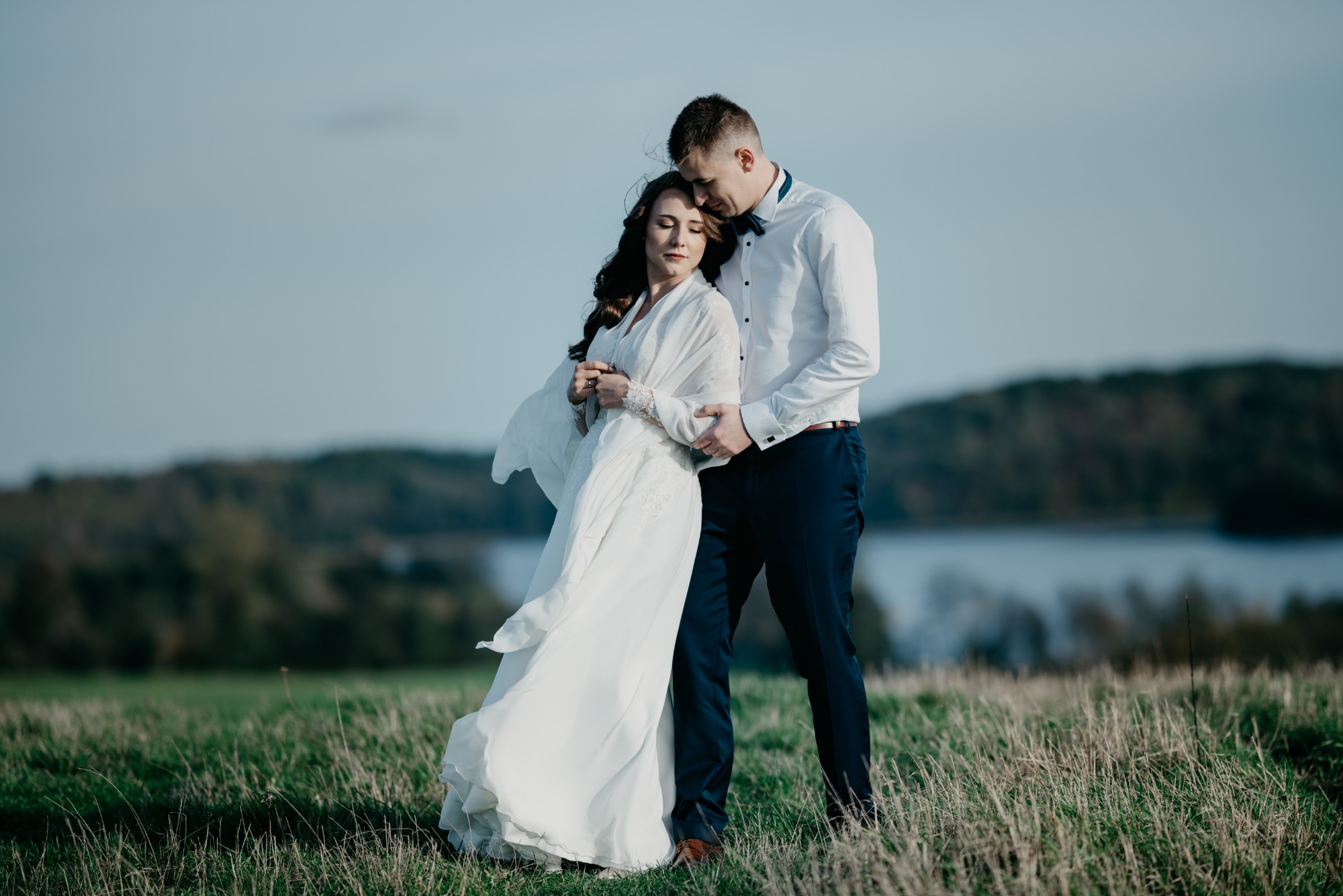 Para narzeczonych w objęciach na łące z jeziorem w tle, panna młoda w zwiewnej sukni z koronkowymi rękawami, pan młody w białej koszuli i granatowych spodniach.
