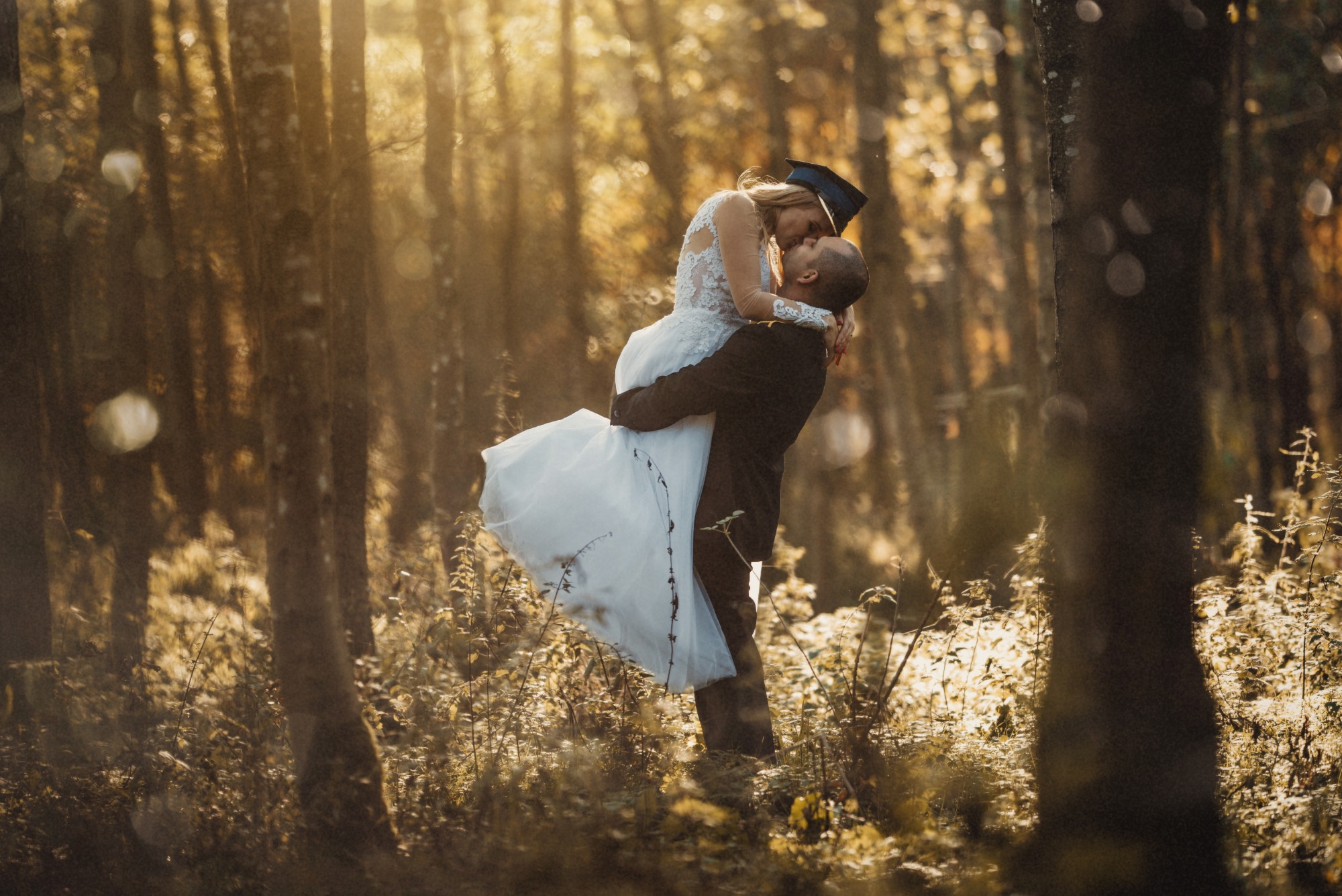 Para w lesie: mężczyzna w ciemnym garniturze trzyma na rękach kobietę w białej sukni i czapce absolwenckiej, całując ją na tle drzew w złotym świetle.