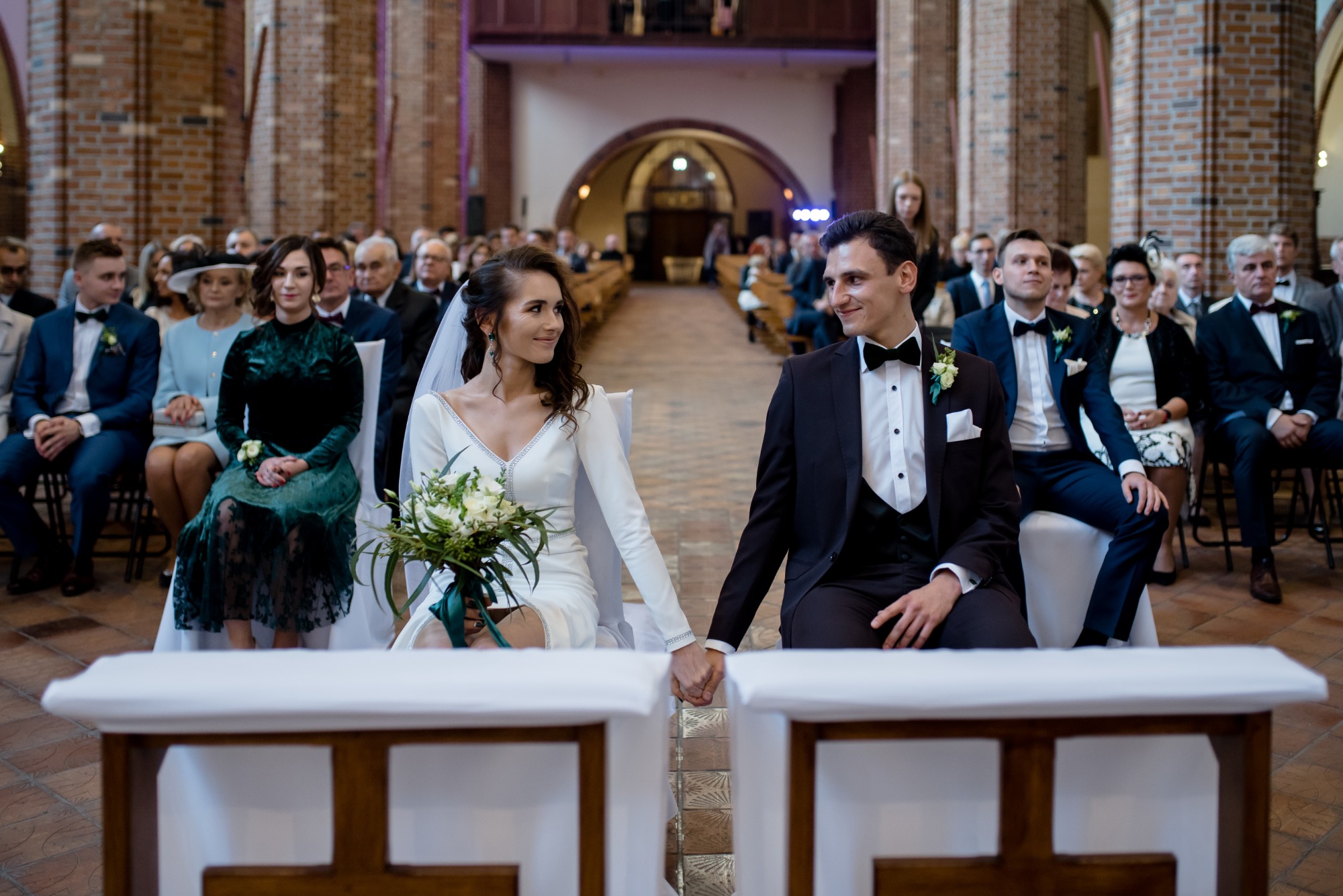 Para młoda trzymająca się za ręce podczas ceremonii ślubnej w kościele, z gośćmi w tle i dekoracją z białego materiału na ławkach.