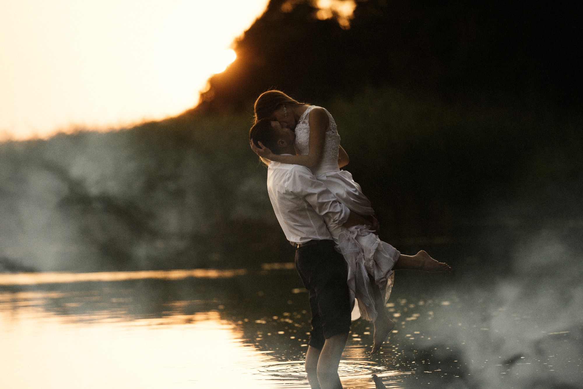 Para młoda w objęciach na tle jeziora o zachodzie słońca, panna młoda w koronkowej sukni jest niesiona przez pana młodego, który stoi w wodzie po kolana.