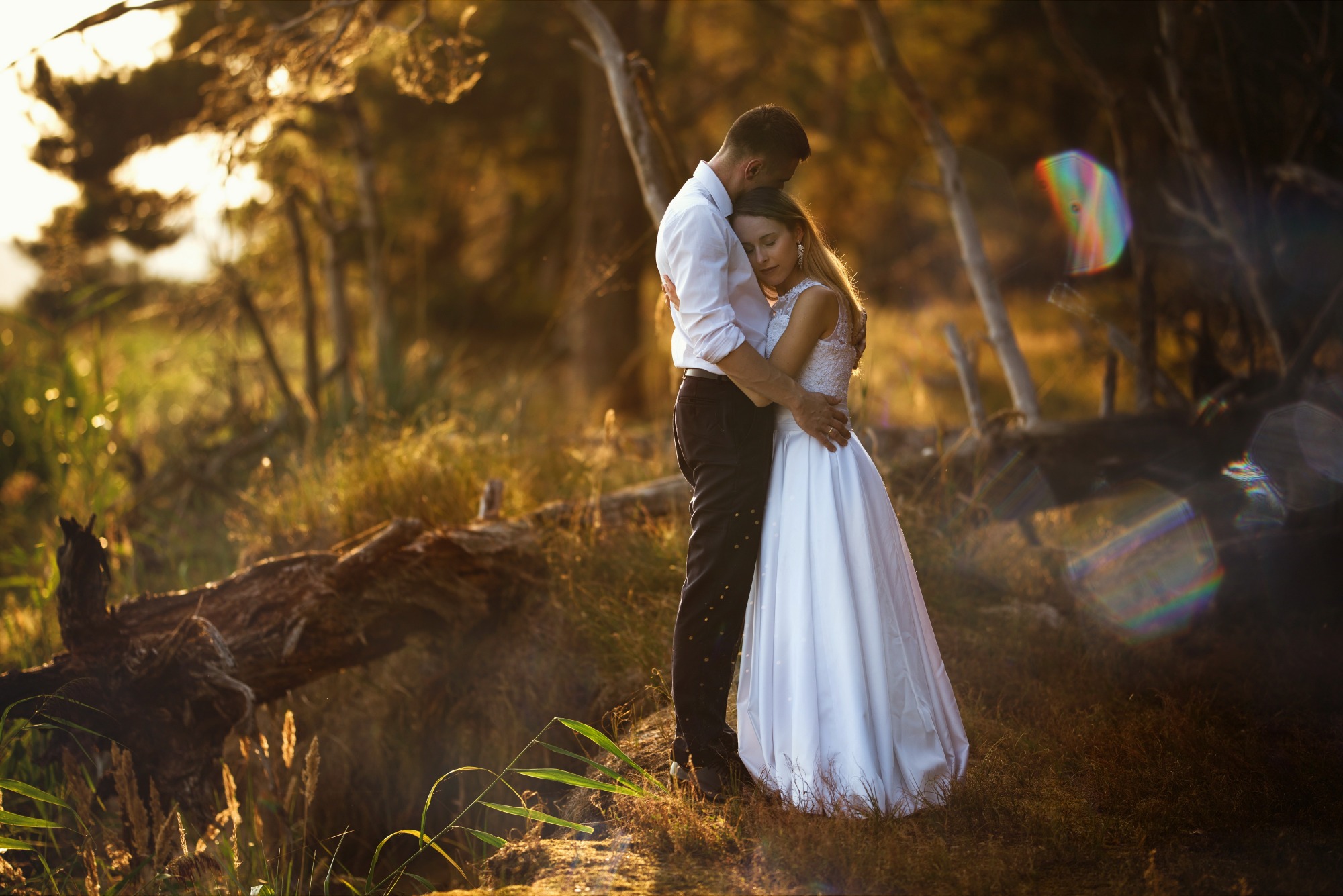 Para narzeczonych w objęciach na tle zalesionego krajobrazu w złotym świetle zachodzącego słońca, ona w białej sukni, on w białej koszuli i ciemnych spodniach.