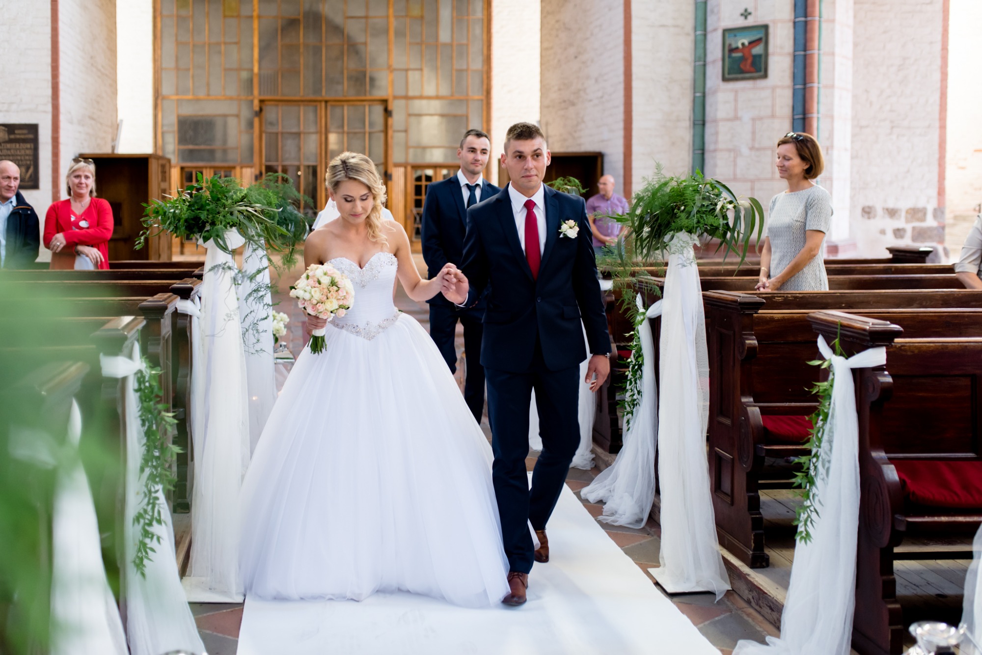 Para młoda, panna młoda w białej sukni z bukietem i pan młody w granatowym garniturze z czerwonym krawatem, idą alejką w kościele, w tle goście.