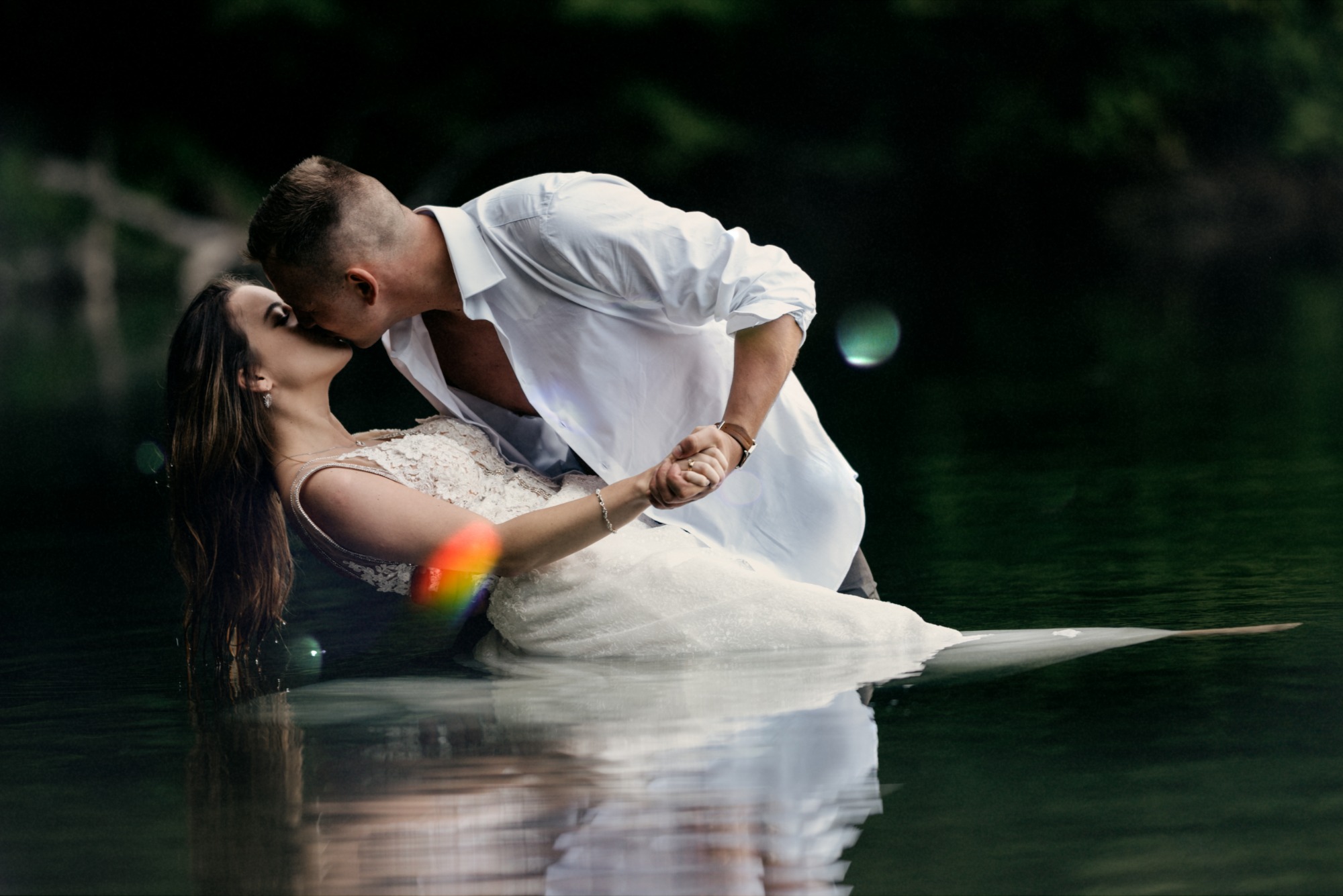 Para młoda w białych strojach, zanurzona w wodzie po pas, w pocałunku; mężczyzna w rozpiętej koszuli obejmuje kobietę w koronkowej sukni, w tle ciemna zieleń drzew.