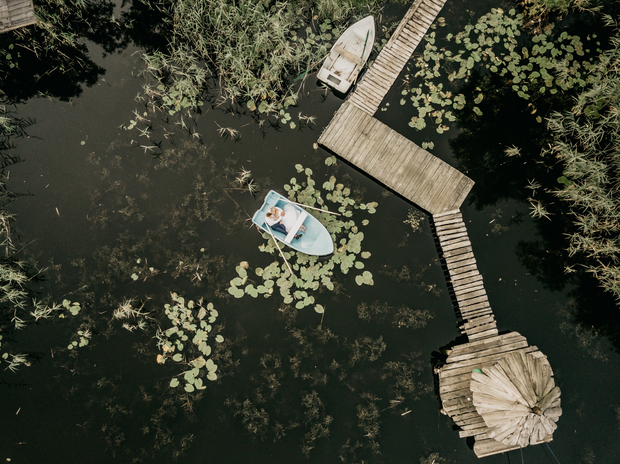 Z lotu ptaka: para w jasnej łódce na ciemnej wodzie z liliami wodnymi, obok drewniane pomosty i druga łódka.