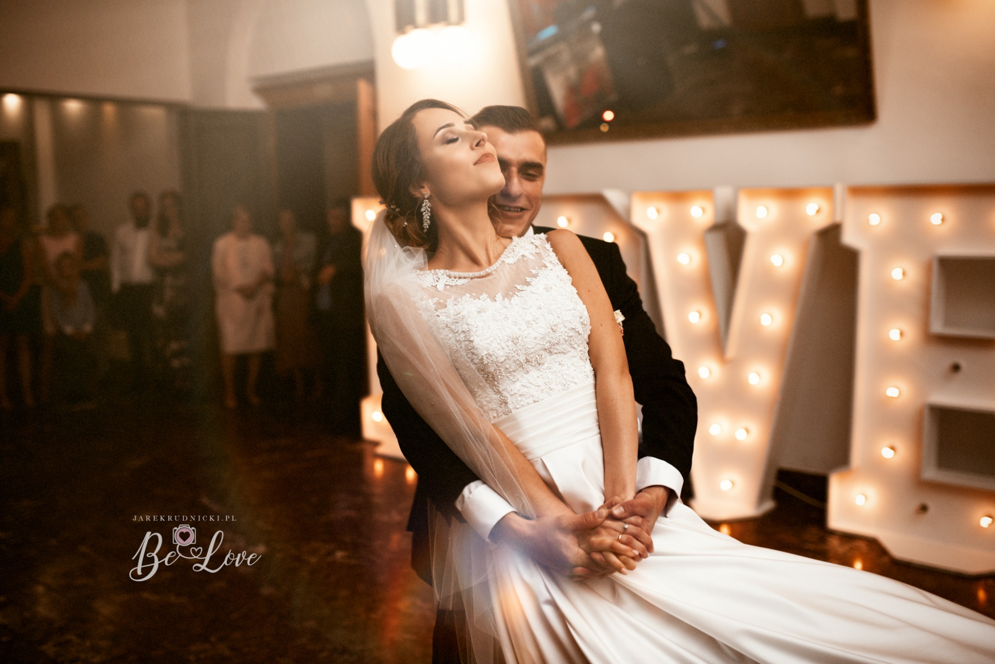 Para młoda w tańcu, panna młoda w białej sukni z koronkową górą i welonem, pan młody w czarnym garniturze, w tle dekoracja LOVE z podświetlanych liter, goście w tle.