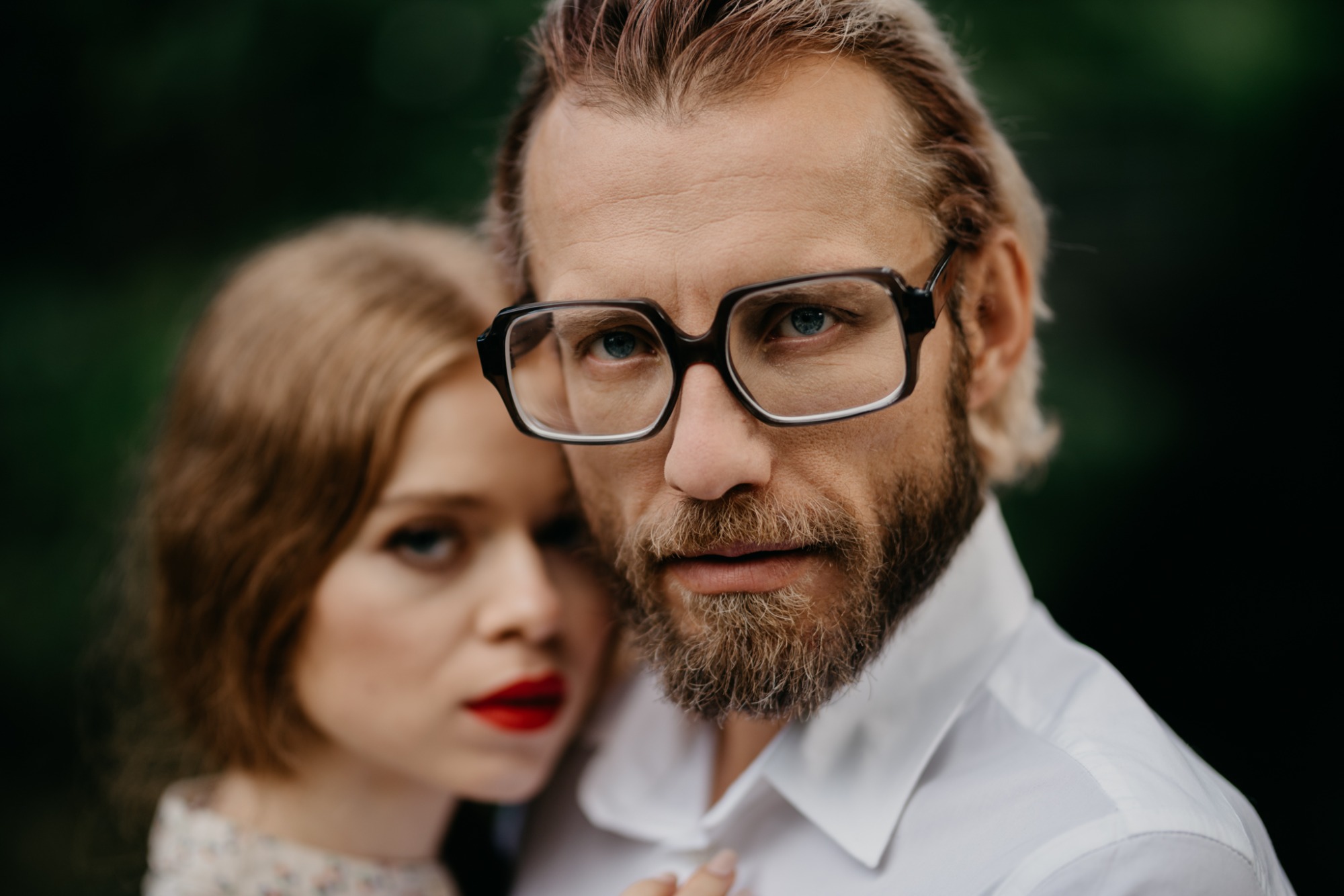 Portret pary: mężczyzna w okularach z bliska, kobieta z czerwonymi ustami w tle.