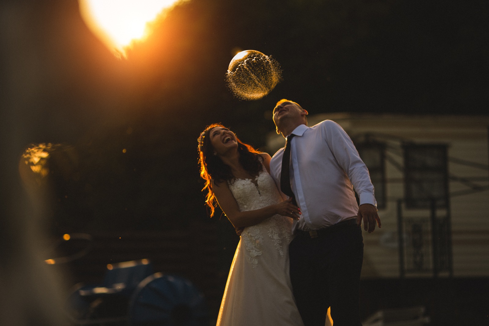 Para młoda, panna młoda w koronkowej sukni i pan młody w białej koszuli i krawacie, śmieją się i patrzą w górę na sferyczną chmurę rozpryskującej się wody, podświetloną zachodzącym słońcem.