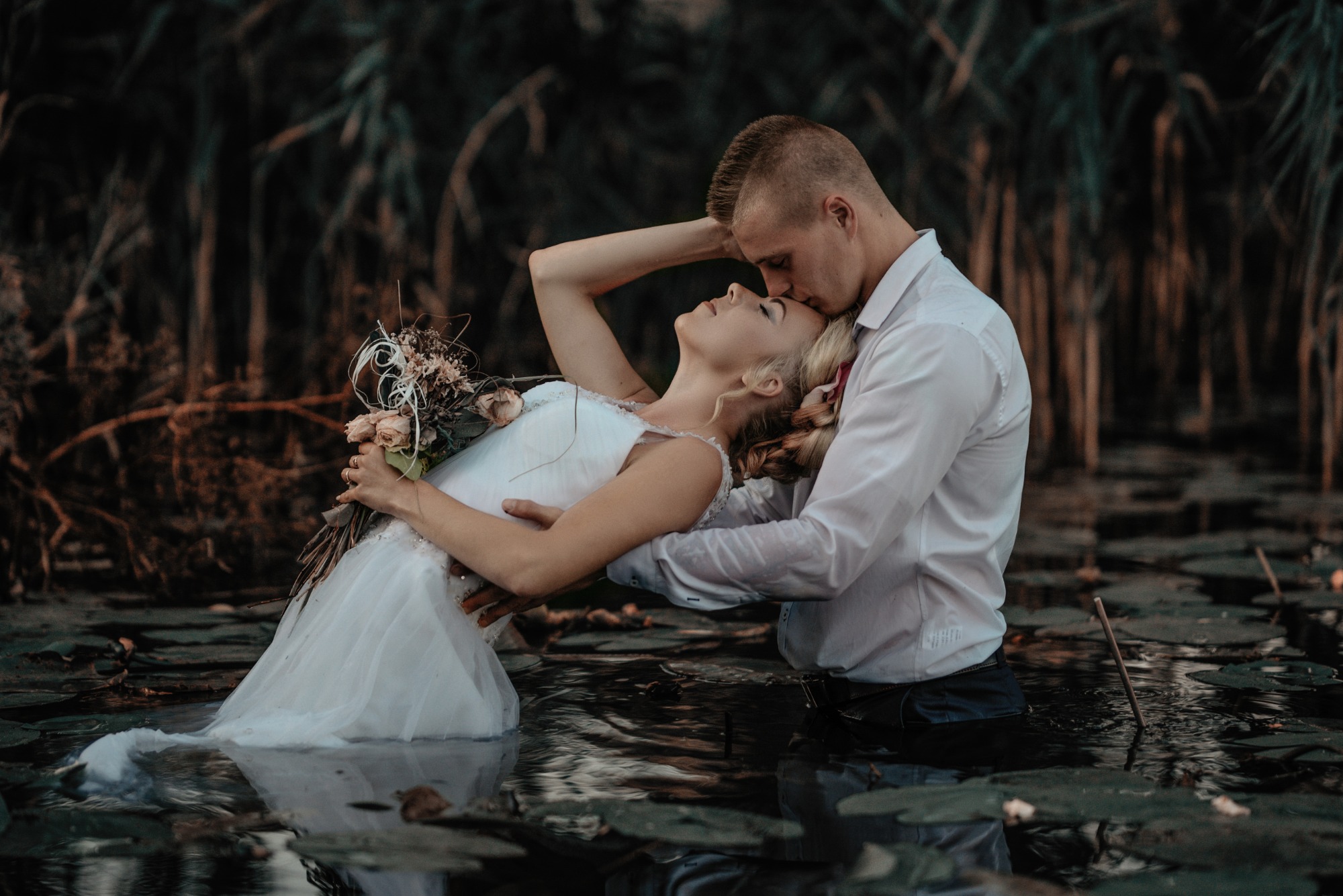 Para młoda w wodzie wśród lilii wodnych: mężczyzna całuje kobietę w czoło, ona trzyma bukiet z suchych kwiatów, romantyczna sceneria.