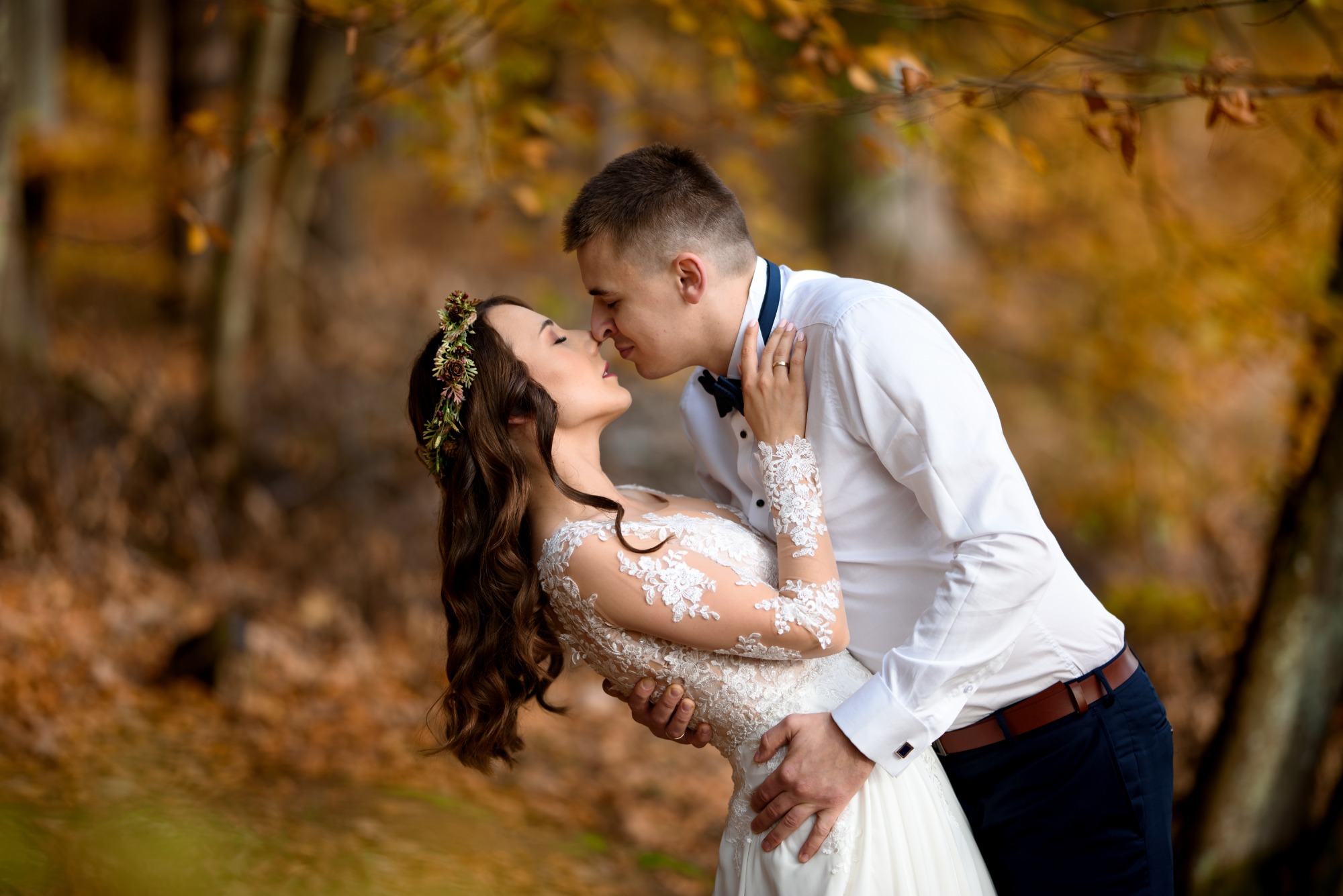 Para młoda w jesiennej scenerii: panna młoda w koronkowej sukni i wianku na głowie, pan młody w białej koszuli i muszce, delikatny pocałunek na tle drzew z żółtymi liśćmi.