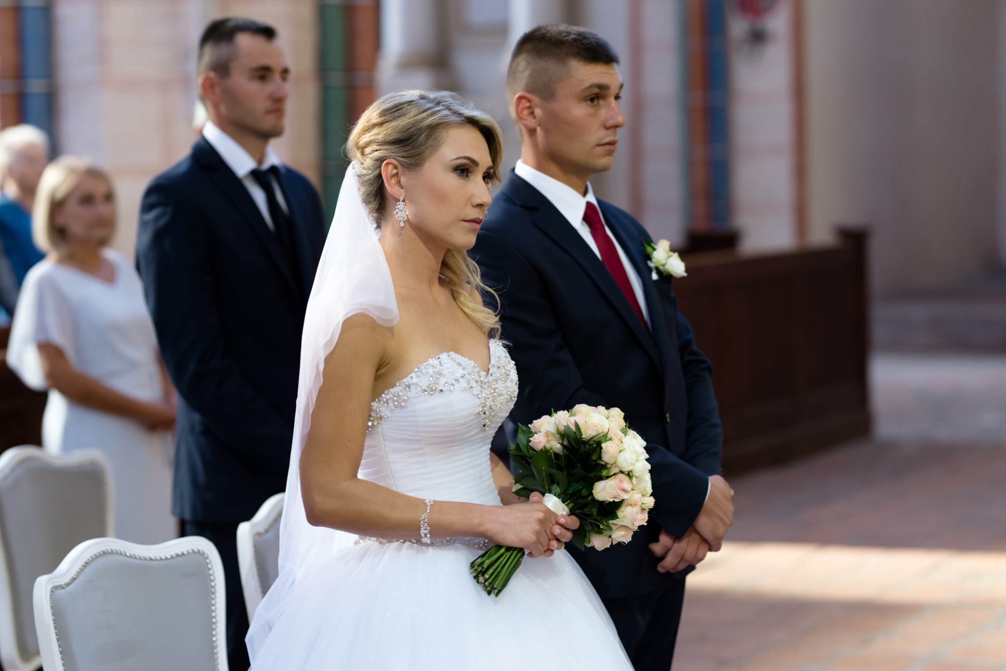 Panna młoda w białej sukni z bukietem białych róż, obok pan młody w granatowym garniturze z czerwoną muchą, w tle goście weselni w kościele.