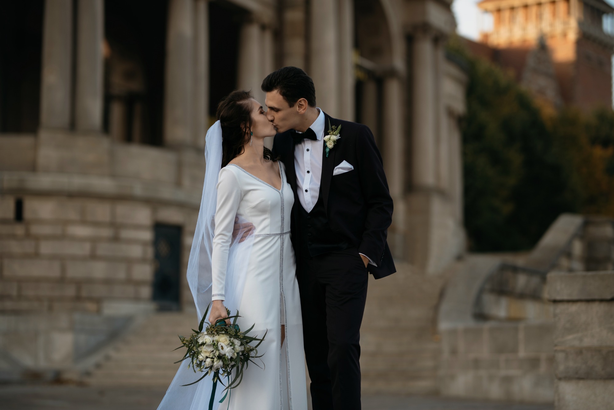 Pocałunek młodej pary w eleganckich strojach ślubnych na tle kolumnady i schodów.