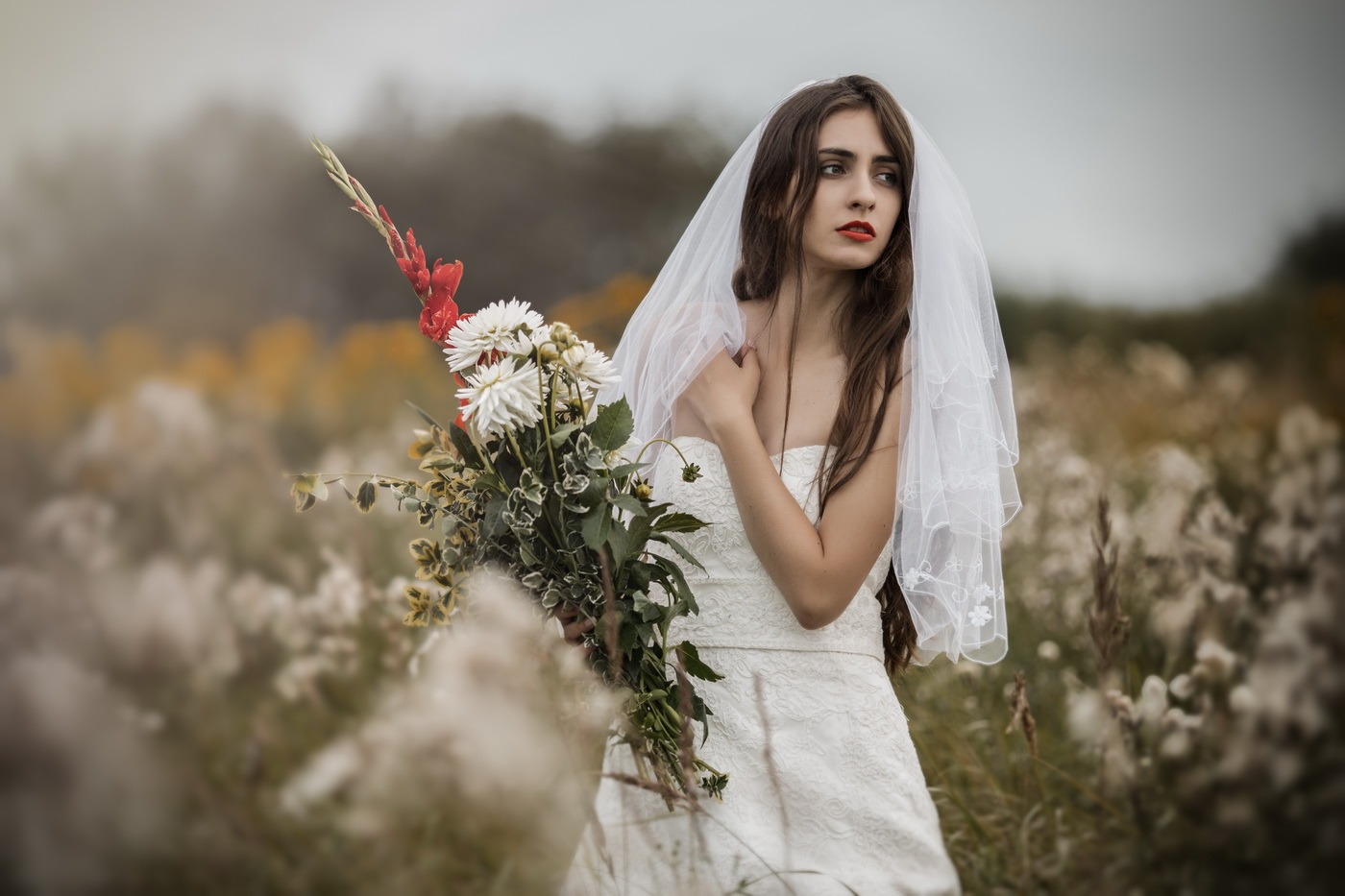 Panna młoda w białej sukni i welonie, trzyma bukiet kwiatów polnych, zamyślona, na tle łąki w jesiennych barwach.