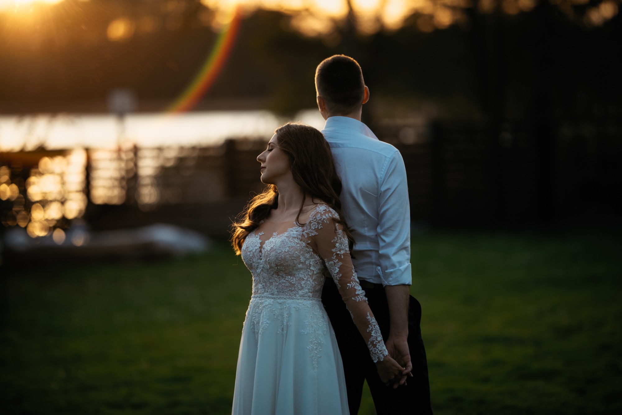 Panna młoda w koronkowej sukni ślubnej opiera się plecami o pana młodego, trzymając go za rękę na tle jeziora o zachodzie słońca z efektem flary.