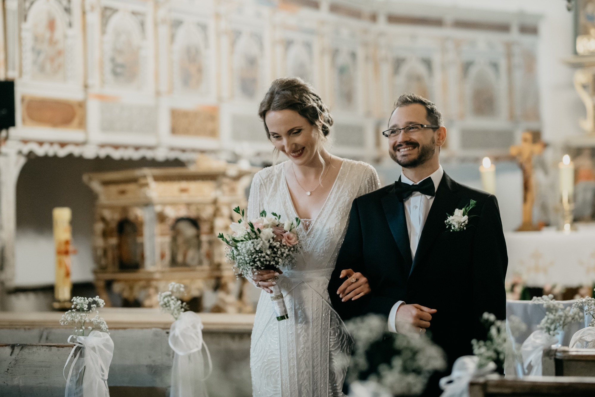 Uśmiechnięta para młoda w kościele: panna młoda w sukni z długim rękawem trzyma bukiet z różowych i białych kwiatów, pan młody w czarnym smokingu z muchą, dekoracje kwiatowe na ławkach.