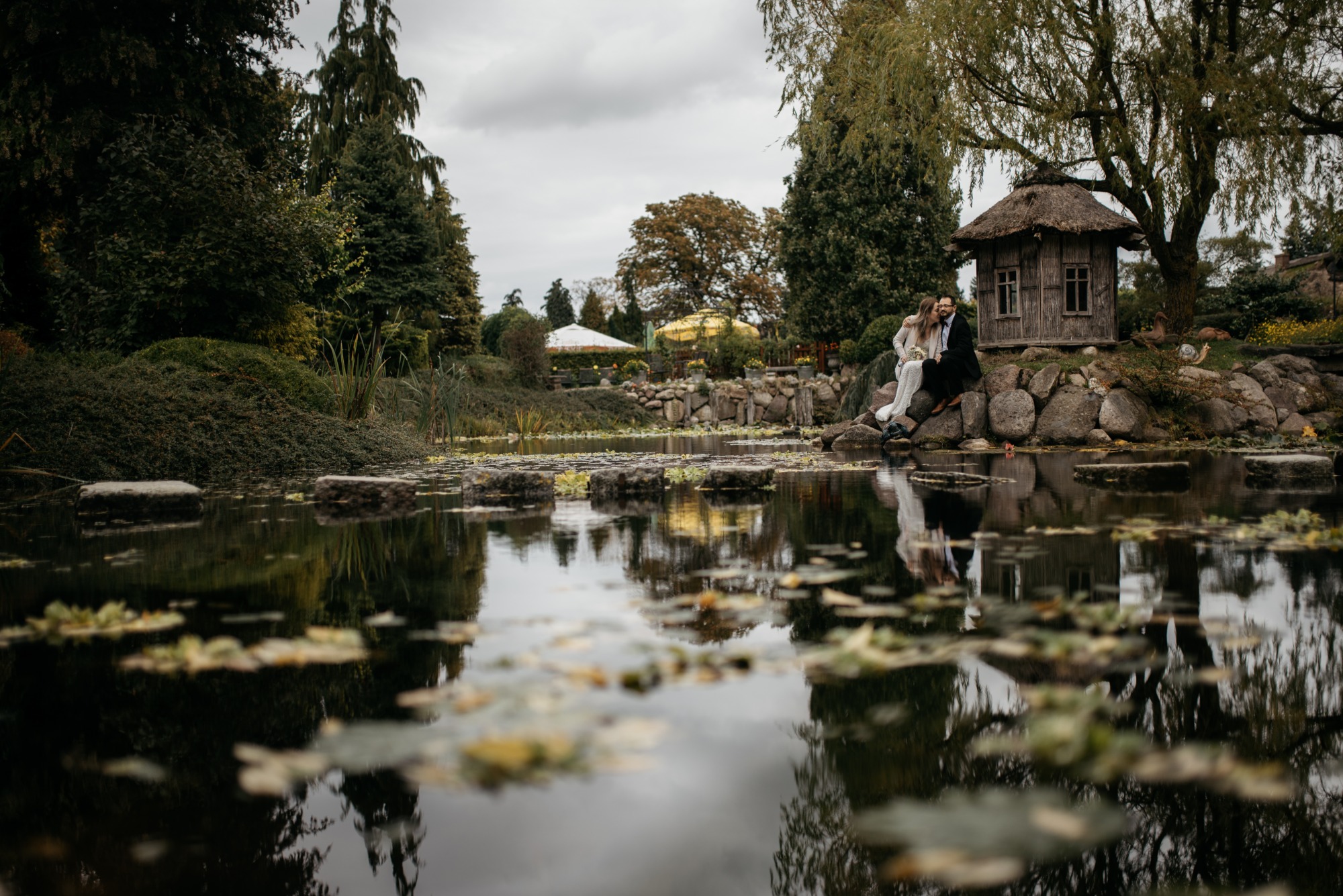 Para młoda siedzi na kamieniach obok stawu z altanką krytą strzechą w tle, odbicie w wodzie.