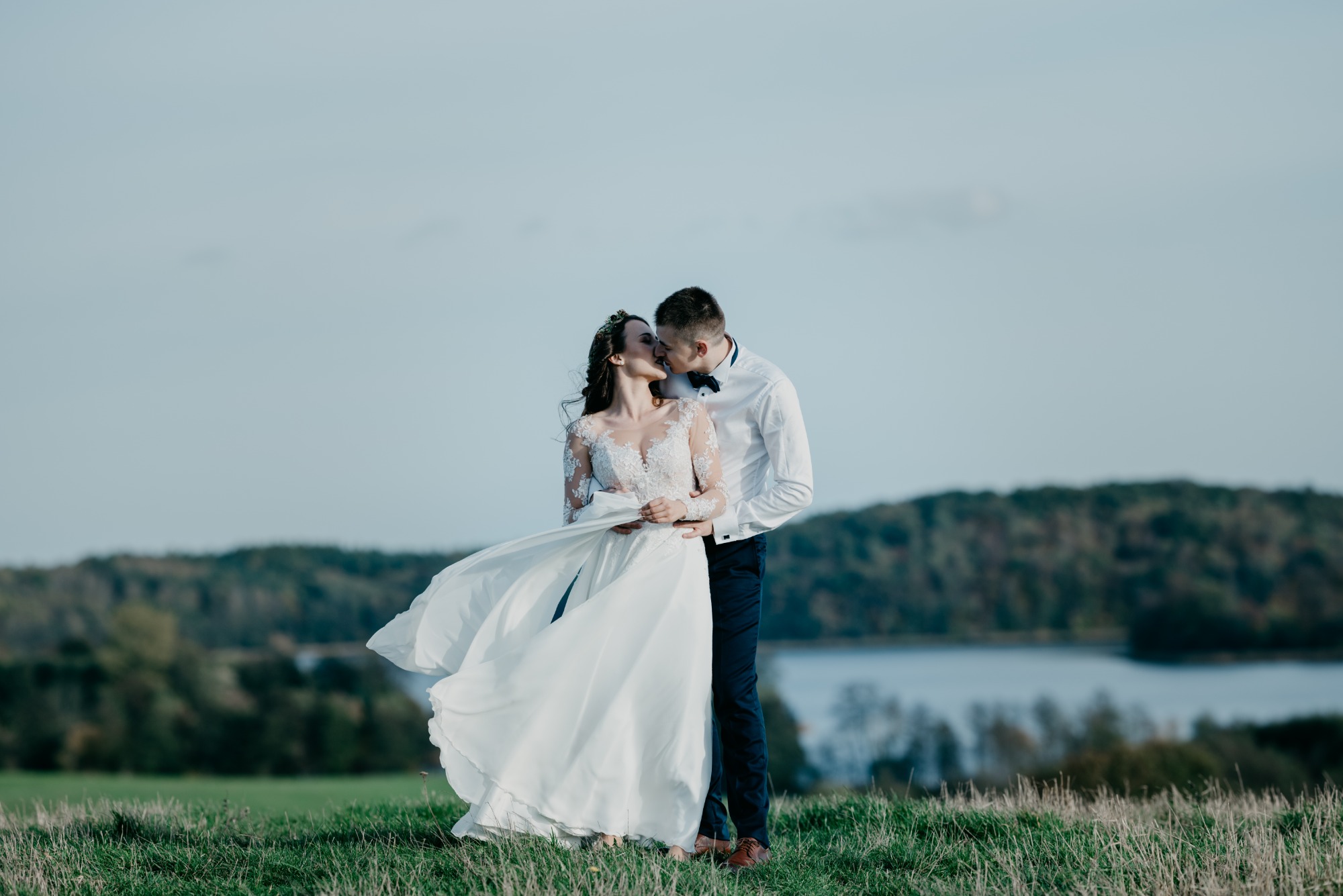 Para młoda całuje się na wzgórzu, suknia powiewa na wietrze, w tle las i jezioro.
