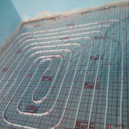 Białe rury ogrzewania podłogowego marki Roth ułożone na siatce z czerwonymi kwadratami, widoczne oznaczenia na podłożu i fragment niebieskiego sufitu z gniazdkiem.